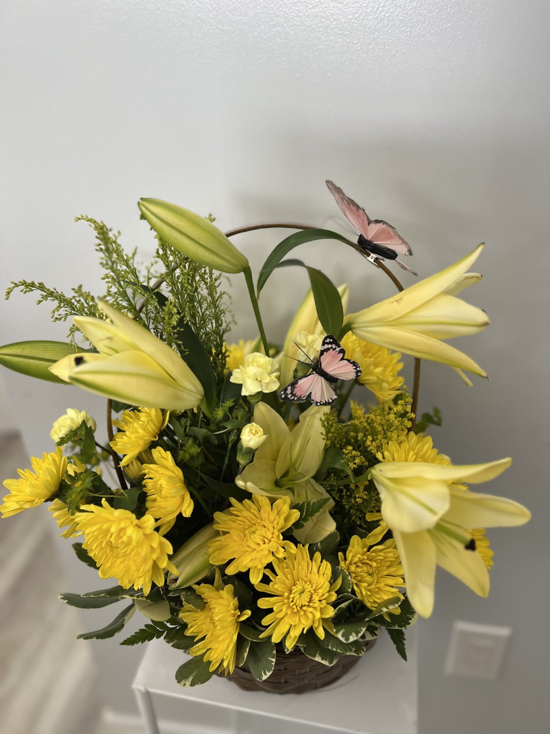 Basket arrangement of yellow lilies and chrysanthemums with pink decorative butterflies.