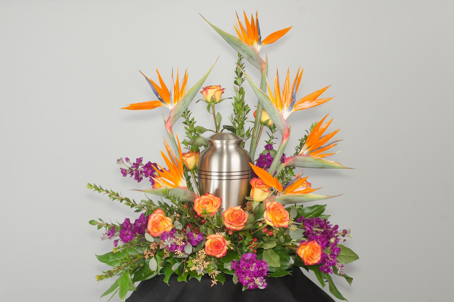 Tropical Sunset - Vibrant and colorful tropical floral ring for the urn of a cherished loved one. This sympathy piece is meant to be as vibrant, colorful and unique as the soul who is being mourned and celebrated. Tropical Birds of Paradise frame the urn and reach for the sky, supported by fluffy orange roses and purple stock saturated in deep color. 