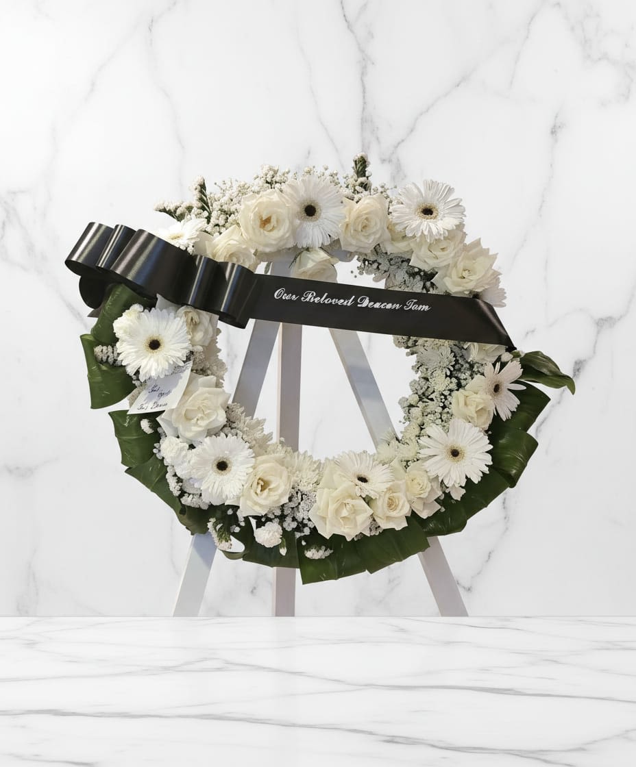 Serenity Circle - A graceful and heartfelt tribute, crafted with fresh white blooms including roses and chrysanthemums, and softly accented with delicate filler flowers and lush greenery, this arrangement expresses sincere condolences. Finished with a classic ribbon with a personalized message from the client. Un homenaje elegante y emotivo, elaborado con frescas flores blancas que incluyen rosas y crisantemos, y suavemente acentuado con delicadas flores de relleno y abundante vegetación; este arreglo expresa condolencias sinceras. Terminado con una cinta clásica con un mensaje personalizado del cliente.