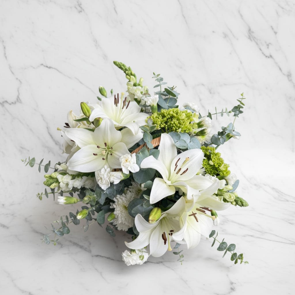 White Serenity Lily Centerpiece - This centerpiece features graceful white lilies surrounded by delicate white chrysanthemum blooms, complemented with fresh eucalyptus leaves and small seasonal filler flowers that add natural texture and softness.  Este centro de mesa presenta elegantes lirios blancos rodeados de delicadas flores de crisantemo blanco, complementados con hojas frescas de eucalipto y pequeñas flores de relleno de temporada que añaden textura natural y suavidad.