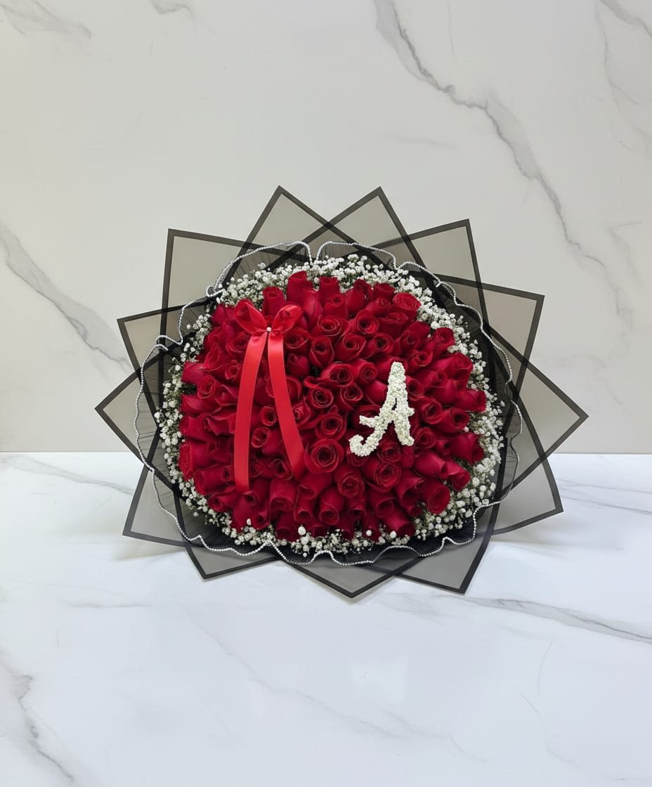 Initial Crimson Roses Bouquet - A beautiful round shaped bouquet made with vibrant red roses, designed to express love, passion, and heartfelt emotion. They are surrounded by delicate gypsophila, and wrapped with black transparent paper for an elegant touch. Un impresionante ramo de rosas rojas intensas dispuestas en un diseño completo y elegante que simboliza el amor, la pasión y el romance eterno. Las vibrantes rosas están bellamente envueltas en papel floral de tonos suaves y neutros, y rematadas con una delicada cinta, creando una presentación refinada y lujosa.