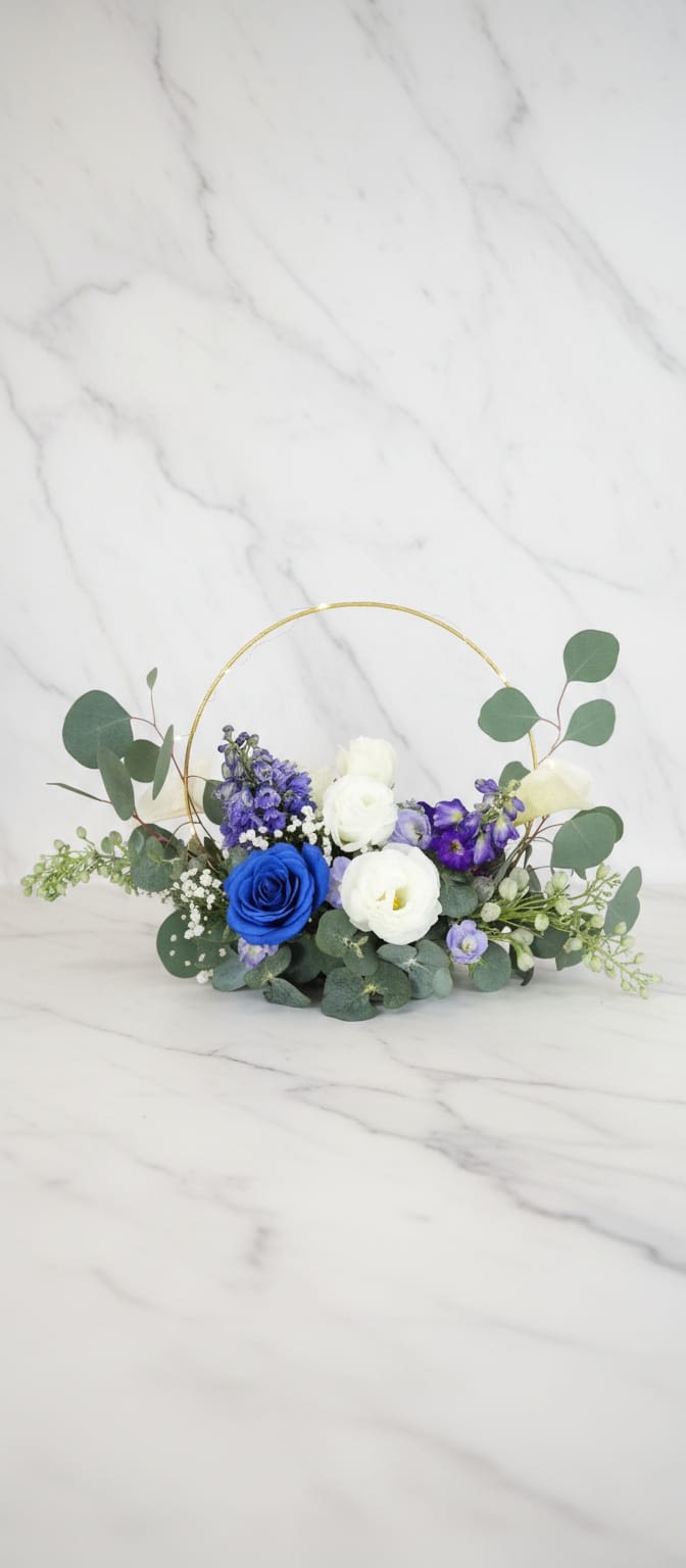 Natural Bloom Harmony - A modern and elegant floral centerpiece featuring a beautiful blend or sprayed blue, white, and soft purple blooms accented with fresh eucalyptus greenery and gypsophila. Designed around a stylish gold hoop frame, this arrangement creates a unique and contemporary display that brings balance, freshness, and sophistication to any table setting. Un centro de mesa floral moderno y elegante que presenta una hermosa mezcla de flores azules pintadas, blancas y púrpuras suaves, acentuadas con verdor fresco de eucalipto y gypsophila. Diseñado alrededor de un elegante marco circular dorado, este arreglo crea una exhibición única y contemporánea que aporta equilibrio, frescura y sofisticación a cualquier mesa.