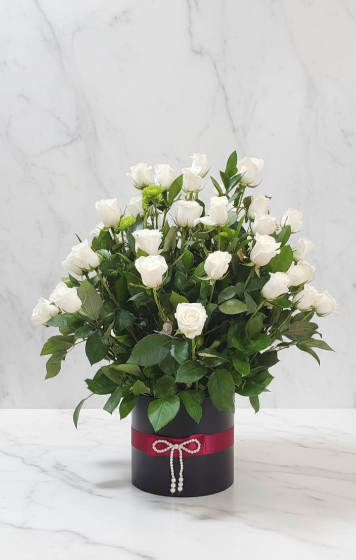 Midnight Ivory Bloom Box - An elegnat and timeless floral arrangement designed to bring softness and sophistication to any space. This beautiful composition features fresh white roses carefully arranged with lush greenery inside a modern black box, finished with a delicate red ribbon. Un elegante y atemporal arreglo floral diseñado para aportar suavidad y sofisticación a cualquier espacio. Esta hermosa composición cuenta con frescas rosas blancas cuidadosamente dispuestas junto con frondoso follaje dentro de una caja negra moderna, rematada con un delicado lazo rojo.