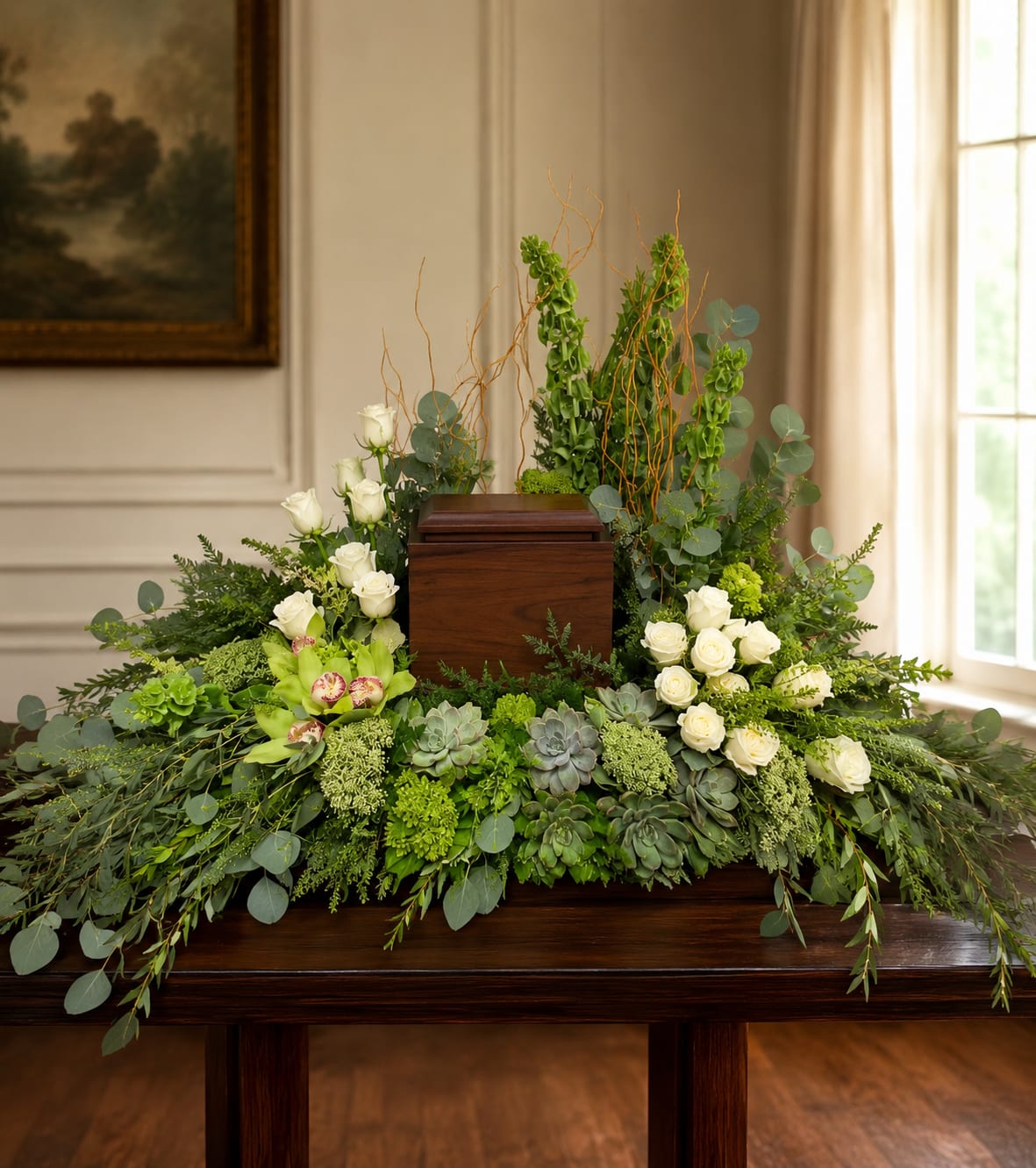Nature‘s Own Cremation Wreath  - A long, low cremation wreath that brings a calm, natural presence to sympathy and funeral settings - the kind of arrangement that helps the person you're honoring feel quietly seen. Clusters of creamy white roses and green cymbidium orchids form the soft focal band, while succulents and textured green blooms weave a layered, organic look that reads both gentle and sculptural.  Taller vertical stems and curly branches rise at one end to create a modern, asymmetrical silhouette that works well as a memorial centerpiece or a subdued statement on a console or chapel table. The green, white, and soft pink palette keeps the design serene and versatile; the mix of smooth orchid petals, velvety roses, and the fleshy texture of succulents gives visual interest up close and from a distance.  Arrives ready to place with a low profile designed for cremation or tabletop display. For care: keep away from direct heat and replace water as needed for any conditioned stems; succulents are arranged to retain form and require minimal attention. This wreath is a thoughtful way to offer comfort and quiet beauty during a difficult time.