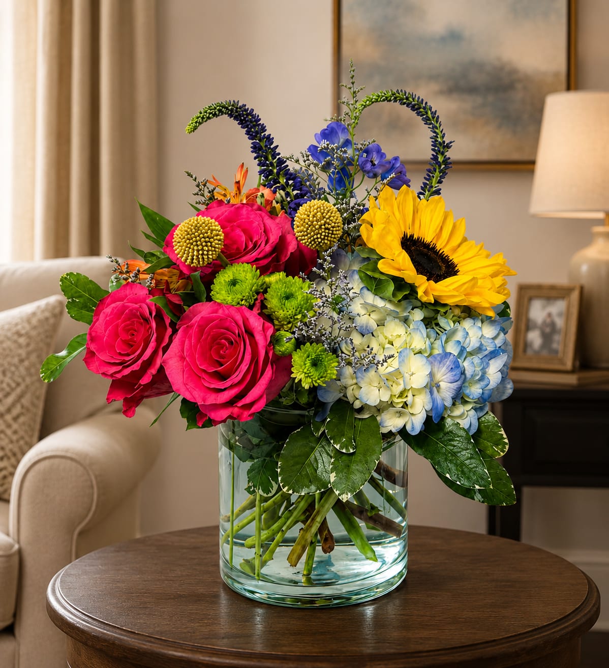 Sumer Sensation By Petal Perfection For The Tampa Bay - A burst of sunny color greets the recipient with bright pink roses paired with a single large yellow sunflower-perfect for a birthday bouquet, congratulations arrangement, or a cheerful everyday gift. This flower arrangement for Tampa Bay delivers a lively, garden-style look that will make the person you're thinking of feel celebrated and noticed the moment it arrives. With delivery all over the Tampa Bay Area. Including Tampa, St Pete, Bradenton Anna Maria and points in between.  Pale blue hydrangea and small green button chrysanthemums soften the contrast and add lush volume, while tall blue‑purple accent spikes lift the composition for graceful height and movement. Arranged in a clear cylindrical glass vase, the mix of warm and cool tones reads joyful and fresh from every angle. Sized to sit beautifully on a dining table, desk, or entry console, it's an upbeat, thoughtful way to send thanks, best wishes, or just-because happiness.