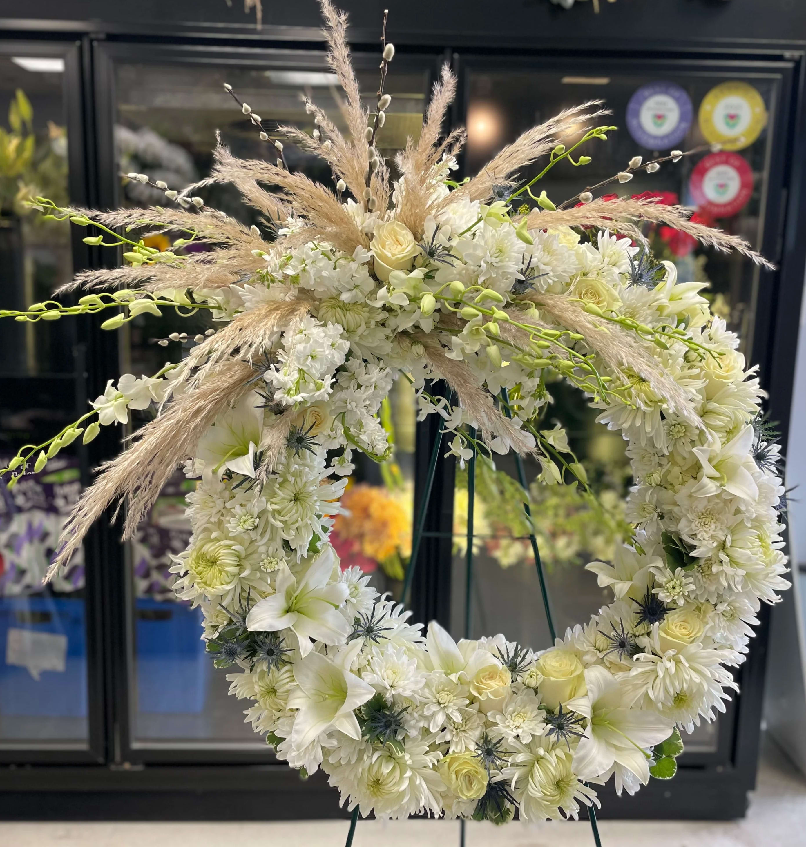 Always Remembered - This large classic wreath is delivered on an easel, and is a thoughtful expression of sympathy and admiration. A standing wreath created from fresh white flowers such as roses, lilies, white orchids, white stock, blue thistles and cushion spray chrysanthemums and accented with greenery. 