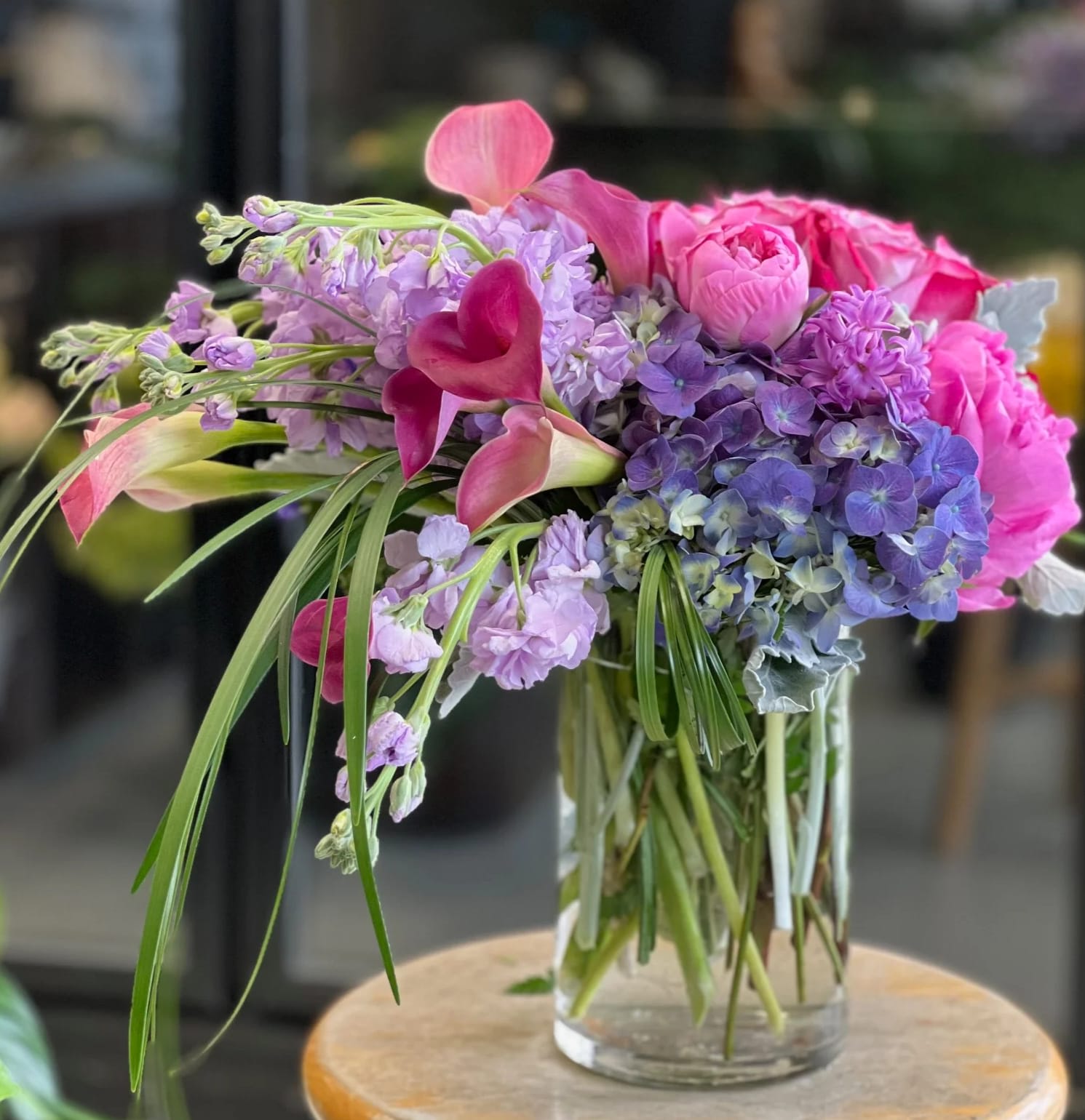 In Her Eyes - Soft, elegant, and full of grace — this exquisite arrangement blends peonies, roses, hydrangeas, calla lilies, stock, dusty miller, and lily grass into a timeless expression of beauty. Designed to capture the depth and warmth found in her gaze, this bouquet is perfect for life’s most meaningful moments.  Please note: Peonies are seasonal and may be thoughtfully substituted with similar blooms of equal value when unavailable.  Two days’ advance notice is required to ensure the freshness and availability of all flowers. This arrangement is not available for same-day delivery or pickup. Feel free to contact us with any questions — thank you for your understanding!