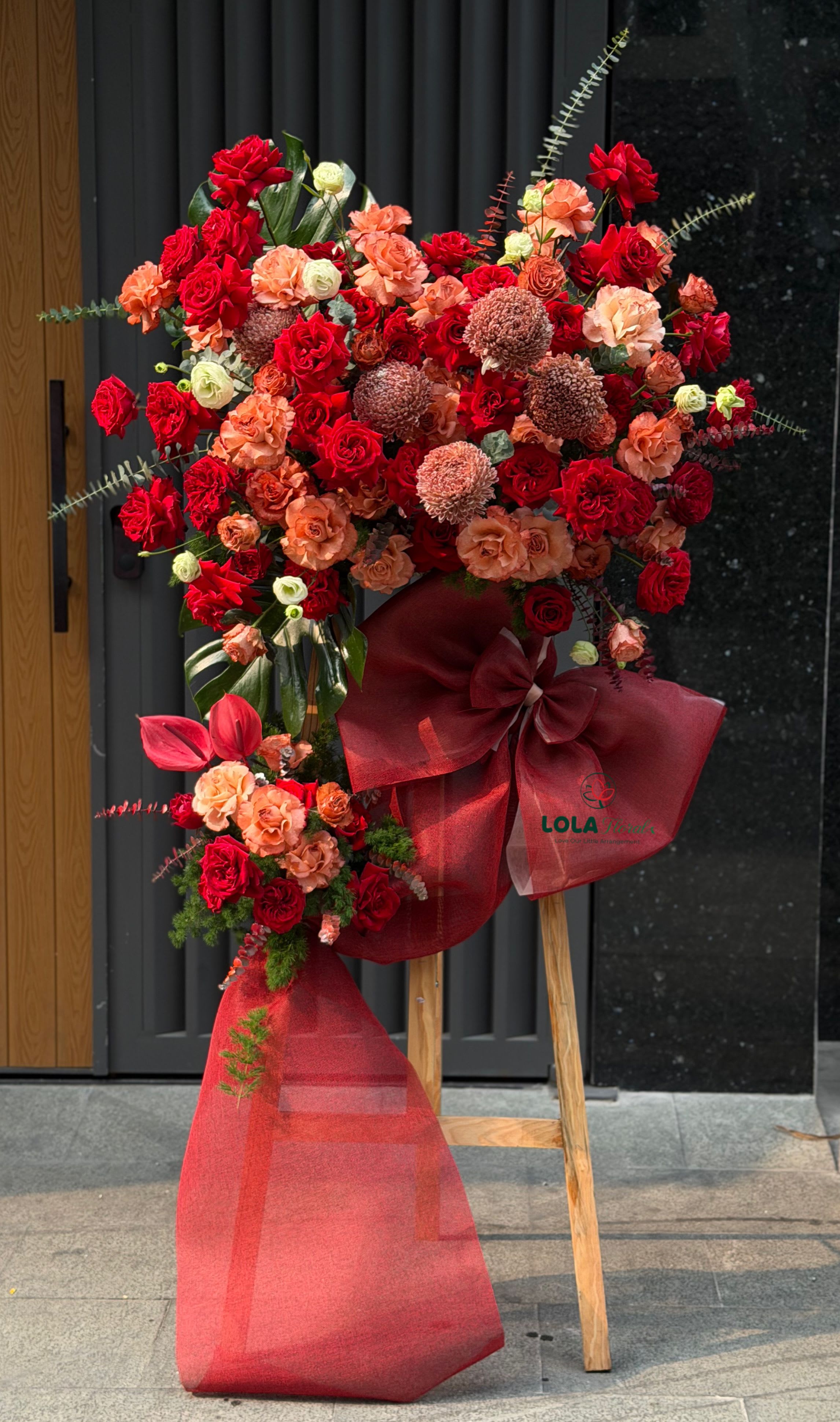 Grand Opening Blush  - A towering display of deep red roses and warm peach blooms-the Velvet Bloom standing arrangement makes an immediate, celebratory statement for a milestone, congratulations, or a romantic occasion. This tall funeral-style or celebratory easel arrangement is built to honor big moments and will make the person you're thinking of feel publicly seen and celebrated.  Dense clusters of red roses, coral spray roses, peach carnations and round white-cream chrysanthemums form a lush vertical composition with tropical foliage peeking through for texture and height. The flowers sit on a sturdy wooden easel and the base is wrapped in a large red sheer fabric with an oversized red decorative bow that echoes the bouquet's rich palette. The arrangement's scale and bold color contrast are designed to read beautifully from a distance.  Ideal for graduations, grand openings, anniversary celebrations, or to mark an important achievement, Velvet Bloom conveys warmth and admiration with a dramatic, gallery-worthy presence. Delivered fully assembled on its easel; light misting and fresh water at the source will keep blooms looking their best-place out of direct sun and away from heat to extend lifespan.