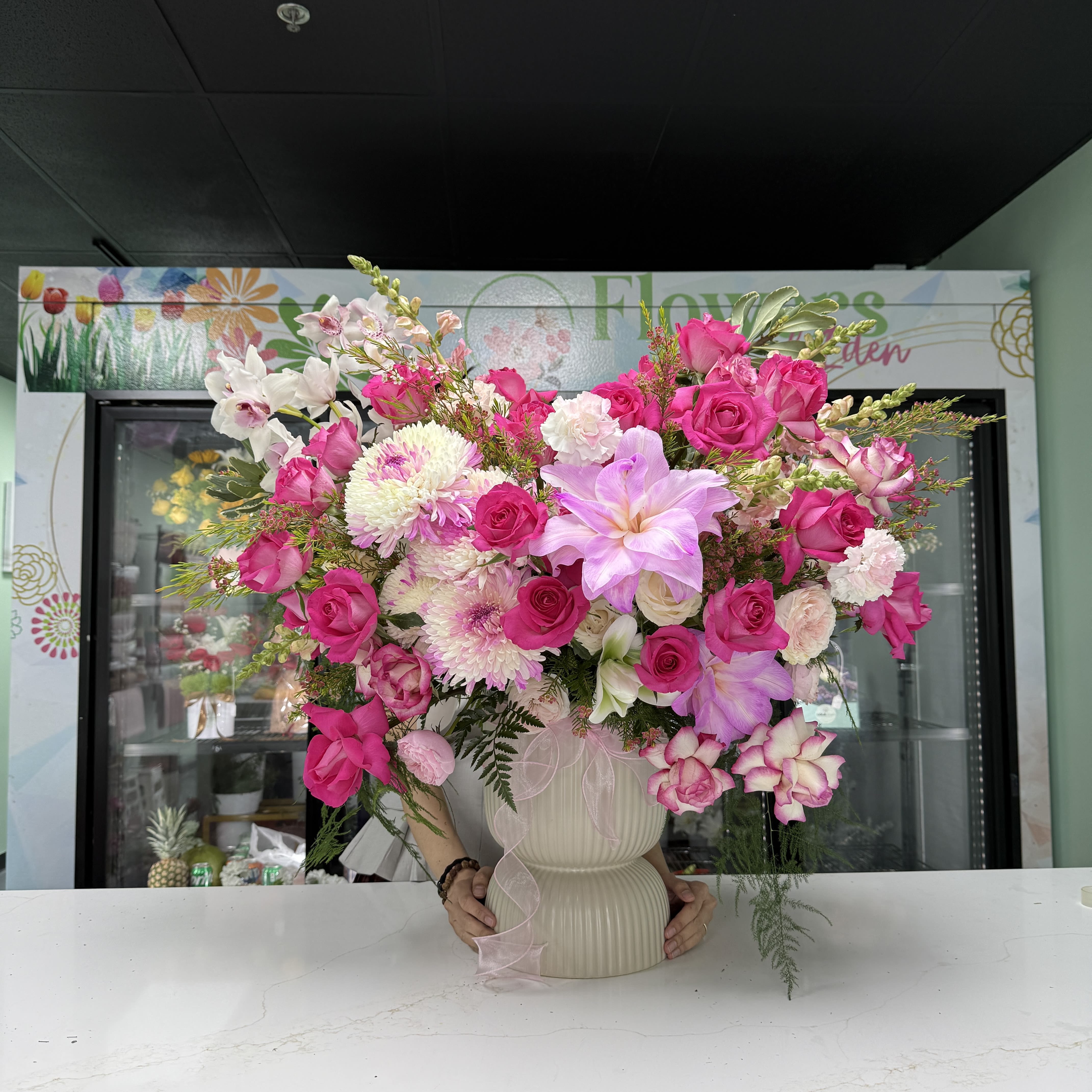 Grand Vase - A lush, overflowing centerpiece of bright pink roses and pale lilies, this flower arrangement is a grand vase bouquet perfect for birthdays, anniversaries, or a thoughtful everyday gift. The wide, celebratory composition feels romantic and cheerful-she'll see the generous sweep of color and know you chose something made to brighten her day.  Built in a ribbed cream ceramic vase, the design layers white chrysanthemums, lavender-toned orchids, and pink carnations with the focal roses and lilies for rich texture and volume. Soft pink ribbon accents tie the palette together; the arrangement is densely packed and spills outward for a full, abundant look that reads as both elegant and joyful.  Ideal for the person you want to make feel truly celebrated, this arrangement suits a dining table, entryway, or office where it can be admired from every angle. The ceramic vase provides a sturdy display - refill water every 2-3 days and trim stems occasionally to keep blooms at their best.