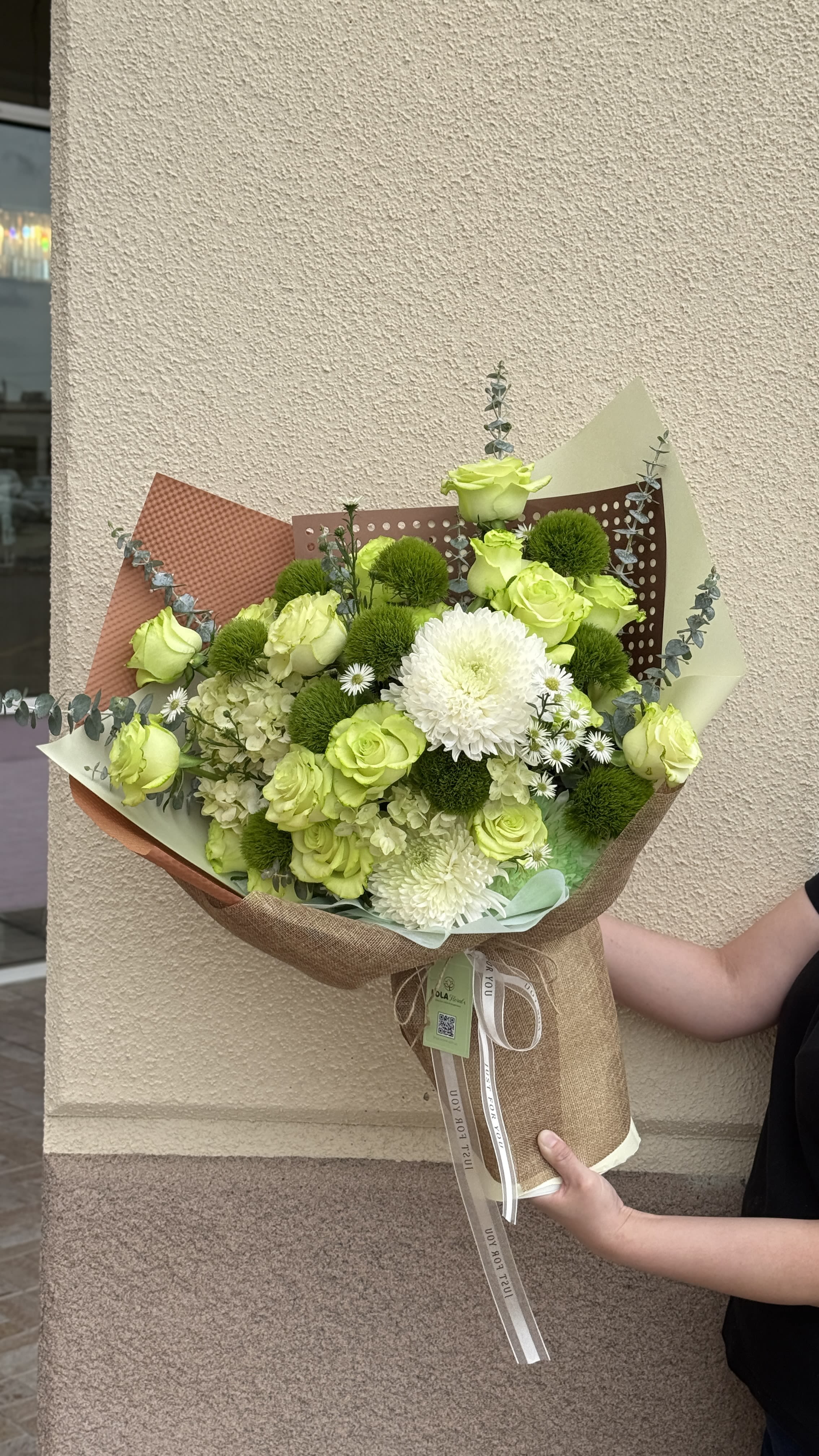 Hand wrapped bouquet 06 - A bright lime-green rose and white chrysanthemum hand-wrapped bouquet that feels both cheerful and slightly formal - perfect as a birthday bouquet, thank-you gift, or to mark a fresh new beginning. The arrangement's vivid green-and-white palette and airy silhouette make the person you're sending it to feel noticed and uplifted the moment it arrives.  This bouquet centers on lime green roses, white chrysanthemums and round green pom-pom blooms with pale hydrangea clusters tucked through for soft volume. Thin eucalyptus-like stems extend beyond the flower mass for a loose, natural outline. It's wrapped in layered brown, cream and decorative perforated paper with burlap accents, finished with a ribbon and hanging tag for a hand-tied presentation; dimensions fall in the classic handheld size suitable for display in a medium vase.  A thoughtful gift for someone who loves modern, fresh florals - ideal for birthdays, congratulations, or "thinking of you" moments. To preserve freshness, trim stems and place in clean water on arrival; if you'd like a different wrap or personalization on the tag, include a note with your order.