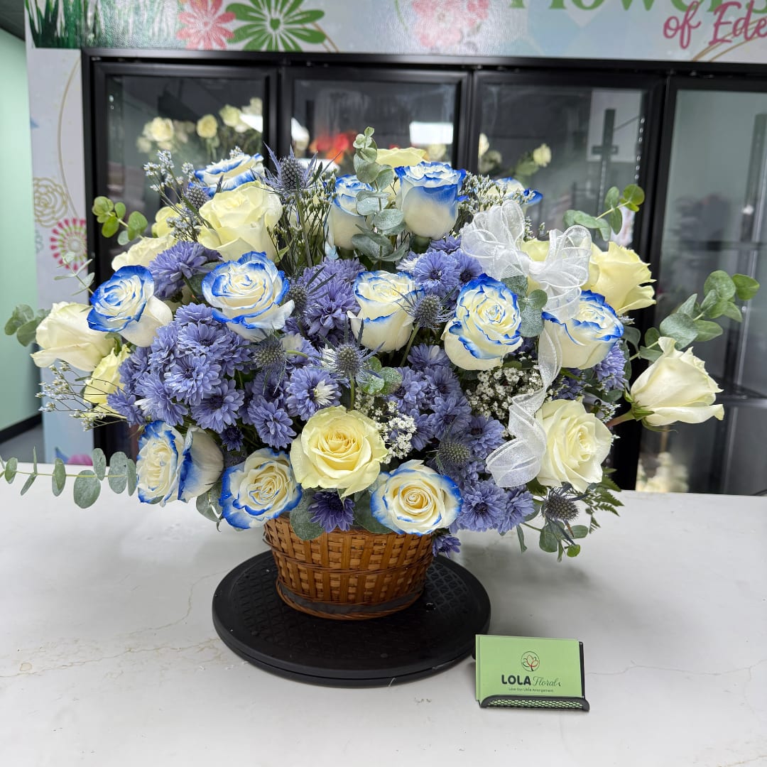 Basket arrangement of blue-edged white roses and purple flowers with a white ribbon