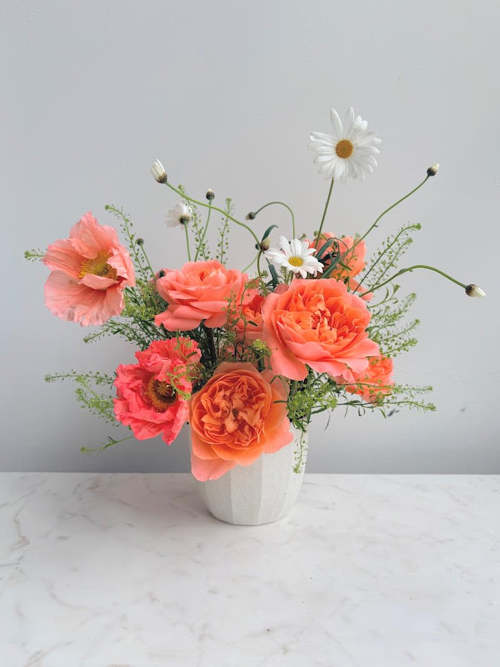 Peach roses and poppy-style blooms with white daisies in a small white ceramic vase