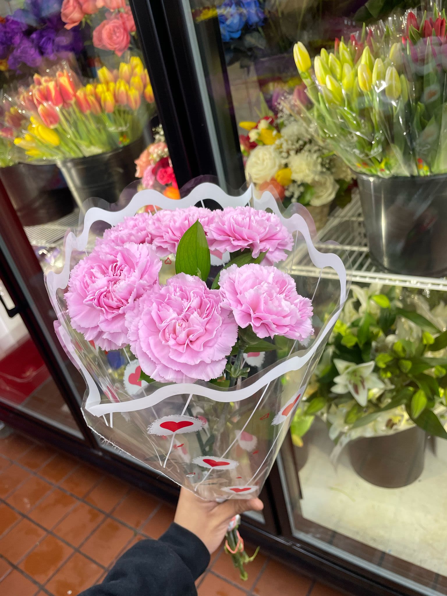 Handheld bouquet of light pink carnations wrapped in a clear sleeve with red hearts.