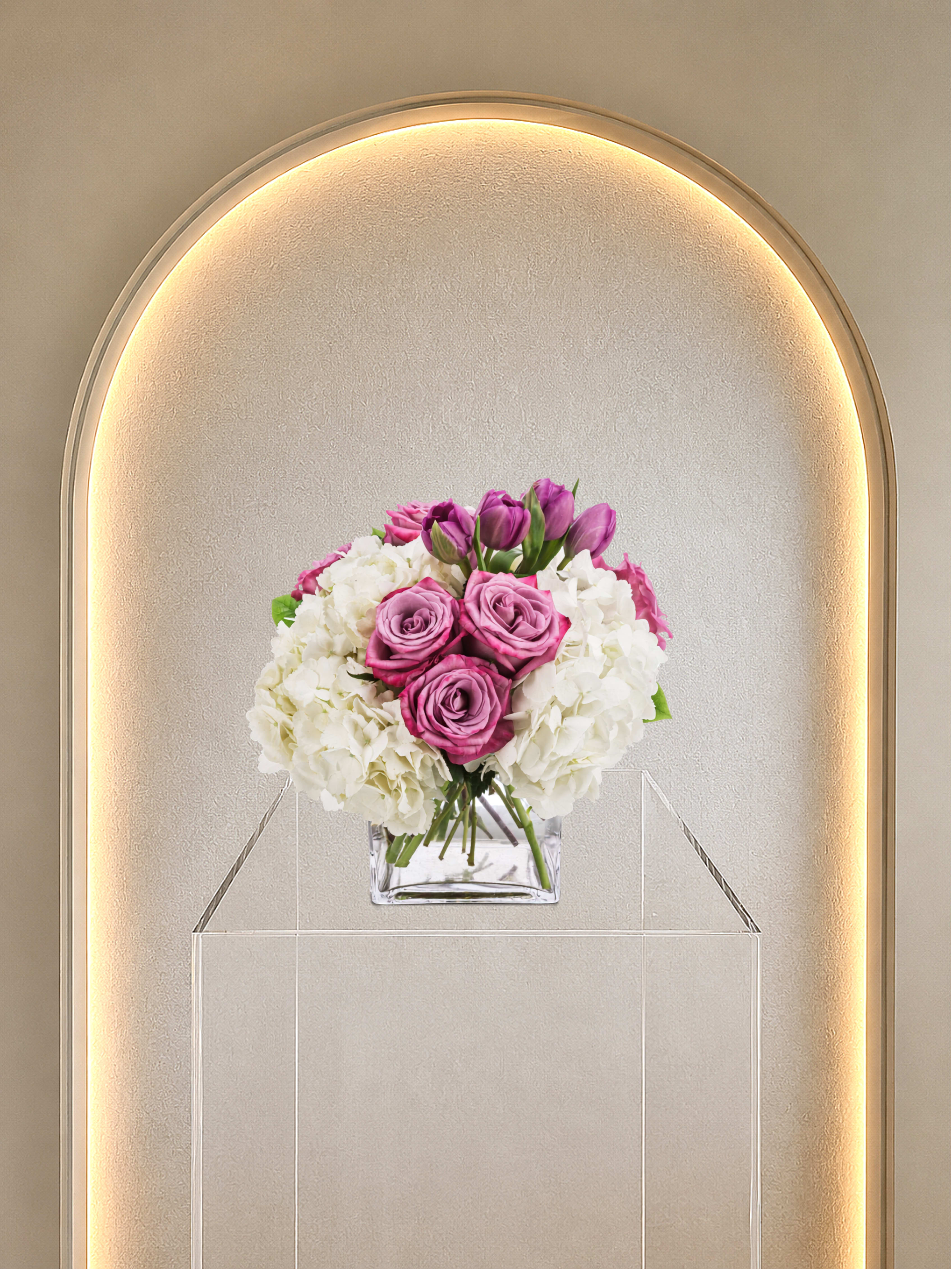 Here For You - Pink roses ring the center of this compact, rounded bouquet, with soft white hydrangea clusters cushioning the sides and a few purple tulips rising above for contrast-an arrangement designed to make the recipient feel seen and comforted. Ideal as a birthday bouquet, anniversary flowers, or a spring celebration arrangement, the mix of pink, white, and purple reads romantic yet modern and sends a clear message of care and thoughtfulness.  Carefully arranged in a clear square glass vase set on a transparent acrylic pedestal, the design has a clean, contemporary presentation that suits both a bedside table and a formal dining setting. Expect lush, full hydrangea blooms, classic pink roses, and upright purple tulips in a tightly rounded silhouette that holds its shape; sizing is compact and polished for easy display. Order this arrangement to give someone a gentle, uplifting surprise-the kind of gift that feels both elegant and reassuring.