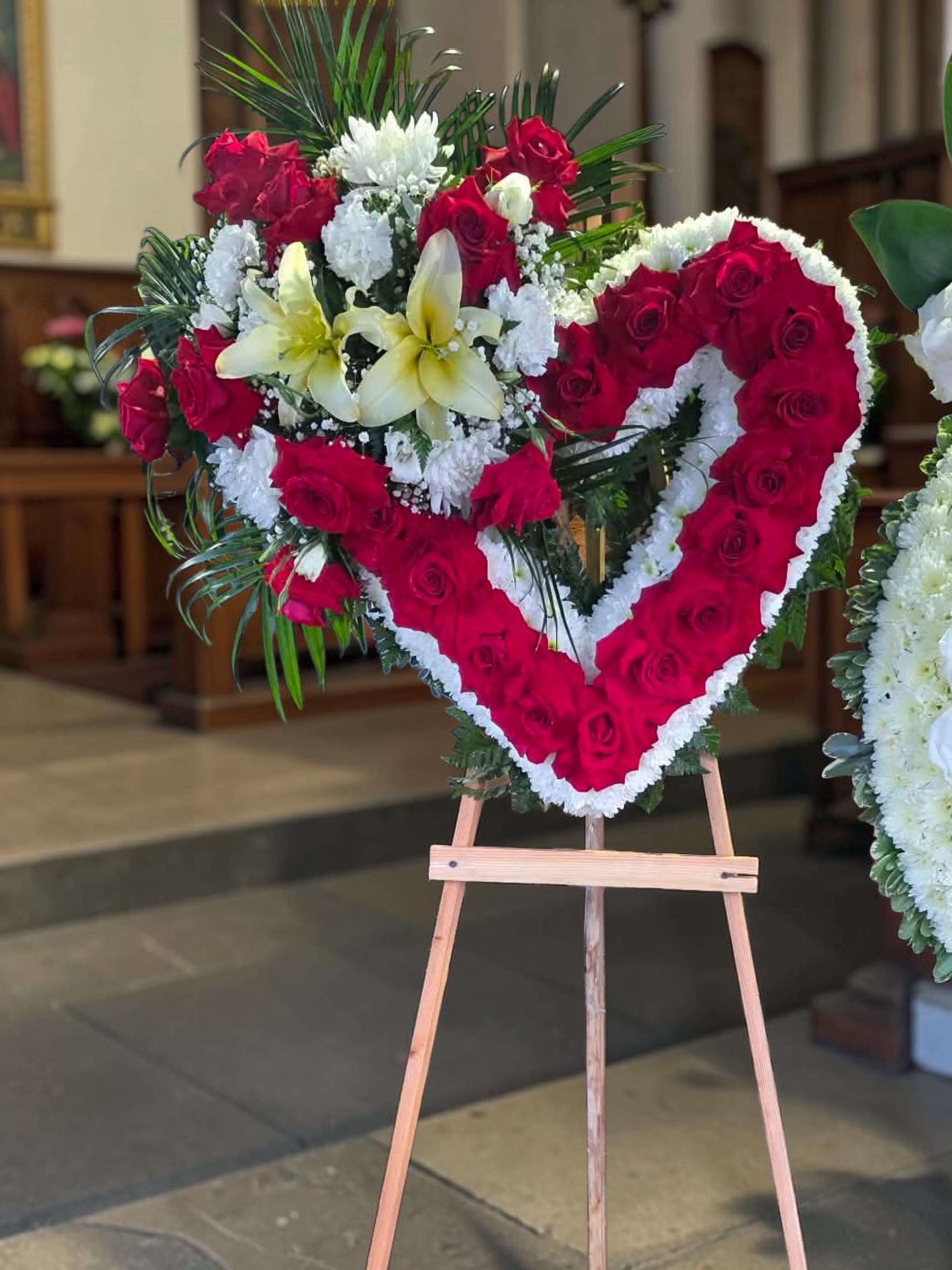 Red Heart love  - A dense heart of deep red roses edged in white blooms creates an immediate, unforgettable statement-this heart-shaped standing tribute is a floral expression of love and remembrance, perfect as a sympathy arrangement or a memorial piece when you want to honor someone dearly loved. The formal red-and-white palette reads ceremonial and heartfelt, while the heart silhouette keeps the message tender and personal for funerals, memorial services, or solemn anniversaries.  Crafted on a sturdy wooden easel, the design pairs the rose heart with a left-side spray of white lilies, additional red roses, yellow accents, green foliage and carnations and daisies for texture and contrast. The mix of roses, lilies, carnations and daisies gives a layered look-velvety rose faces, graceful lily petals, and the soft, ruffled carnations with cheerful daisy highlights-while the green foliage frames and lifts the composition.  Choose this arrangement when you want the recipient's family to feel deeply seen and comforted by a clear symbol of love. It arrives ready to display on its easel for services; please let us know delivery dates and any personalization requests so we can ensure timely placement and a respectful presentation.