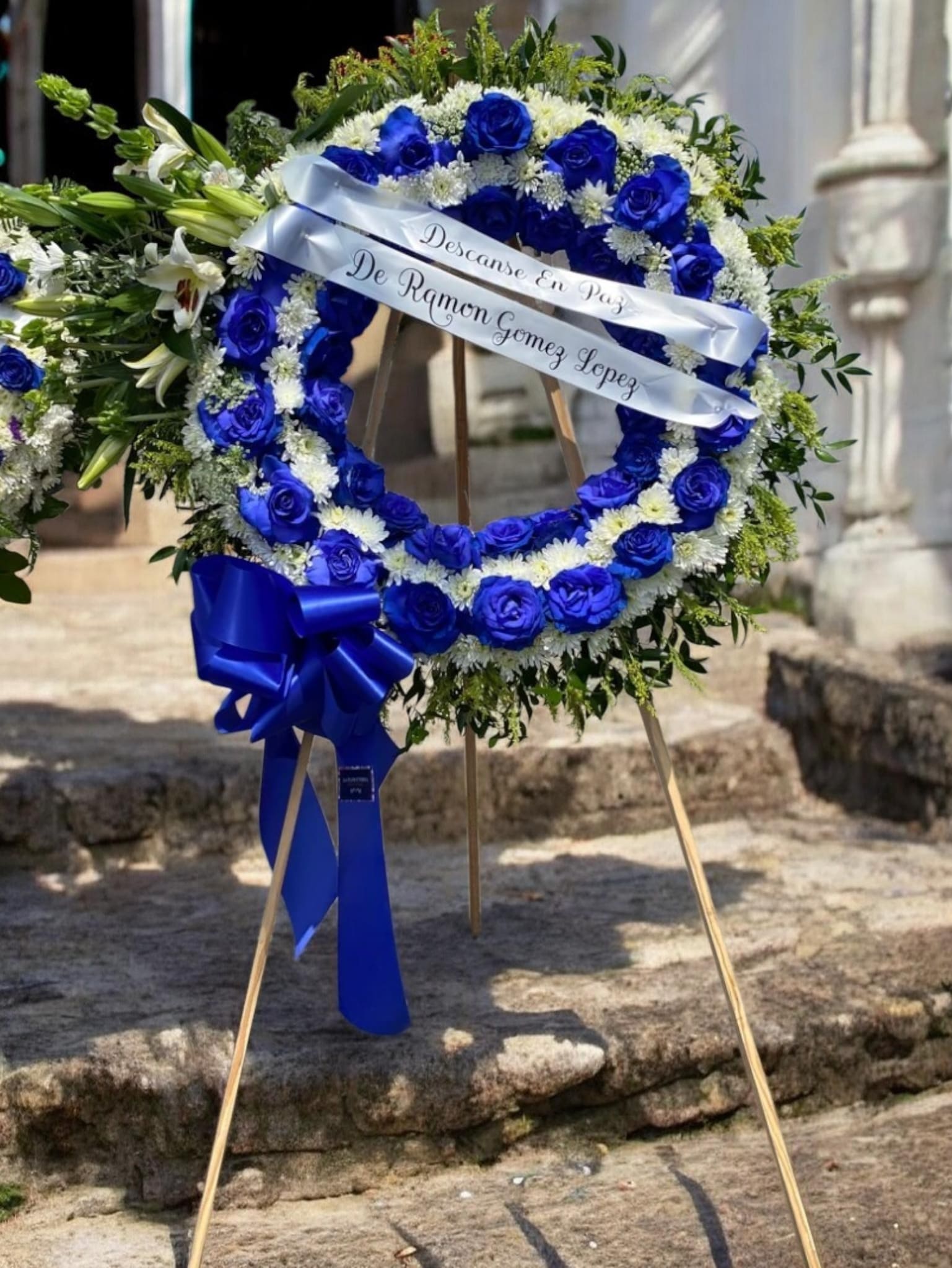 Amor Eterno - Vivid blue roses arranged in a large circular wreath create an immediate, dignified presence-perfect as a funeral wreath or sympathy tribute for a memorial or graveside service. The blue-and-white palette reads formal and comforting, sending a clear message of respect and remembrance to the person you're honoring.  This large circular funeral wreath pairs striking blue roses with white chrysanthemum-like blooms and white lilies, all set against a backdrop of fresh greenery for contrast and texture. It arrives mounted on a floral wreath stand and is finished with a white satin ribbon bearing memorial text across the center and a substantial blue bow near the lower left, giving the tribute a polished, traditional look.  Choose this wreath to express sympathy and to honor a life with grace-the color and composition are designed to be visible and consoling in chapel or graveside settings. If you need custom ribbon wording or delivery timing for a service, include those details at checkout so we can prepare the tribute exactly as requested.