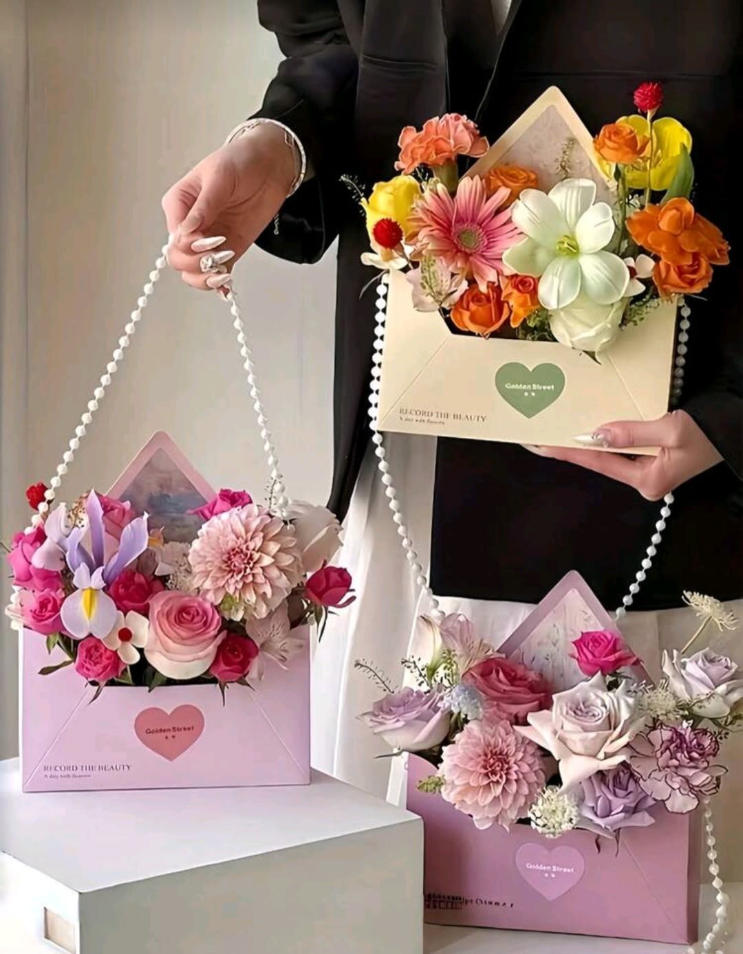 Love Envelope - A mixed flowers make this envelope-style arrangement a delight for birthdays, anniversaries, or a thoughtful "thinking of you" surprise. The presentation reads like a boutique gift: cream and pink envelope boxes with beaded handles, decorative pearl strands, and heart-shaped labels that turn simple blooms into a keepsake moment for the person you're celebrating.  Inside each box a lively, layered mix of roses, gerbera daisies, dahlias, carnations, and small daisy-like fillers fills the space. Colors range from soft blush and lavender roses to bright orange and sunny yellow accents, plus crisp white blooms for contrast-one composition highlights a large pink gerbera and another a pale dahlia for focal texture. Decorative ribbon and pearl details add a polished, feminine touch to the compact, hand-held presentation.  Perfect for sending warm congratulations, a cheerful thank-you, or a Mother's Day surprise, this arrangement makes the recipient feel genuinely seen and celebrated. Boxes arrive ready to gift; the compact clusters are designed for easy handling and display-no vase required-so the flowers look beautiful straight from the envelope box.