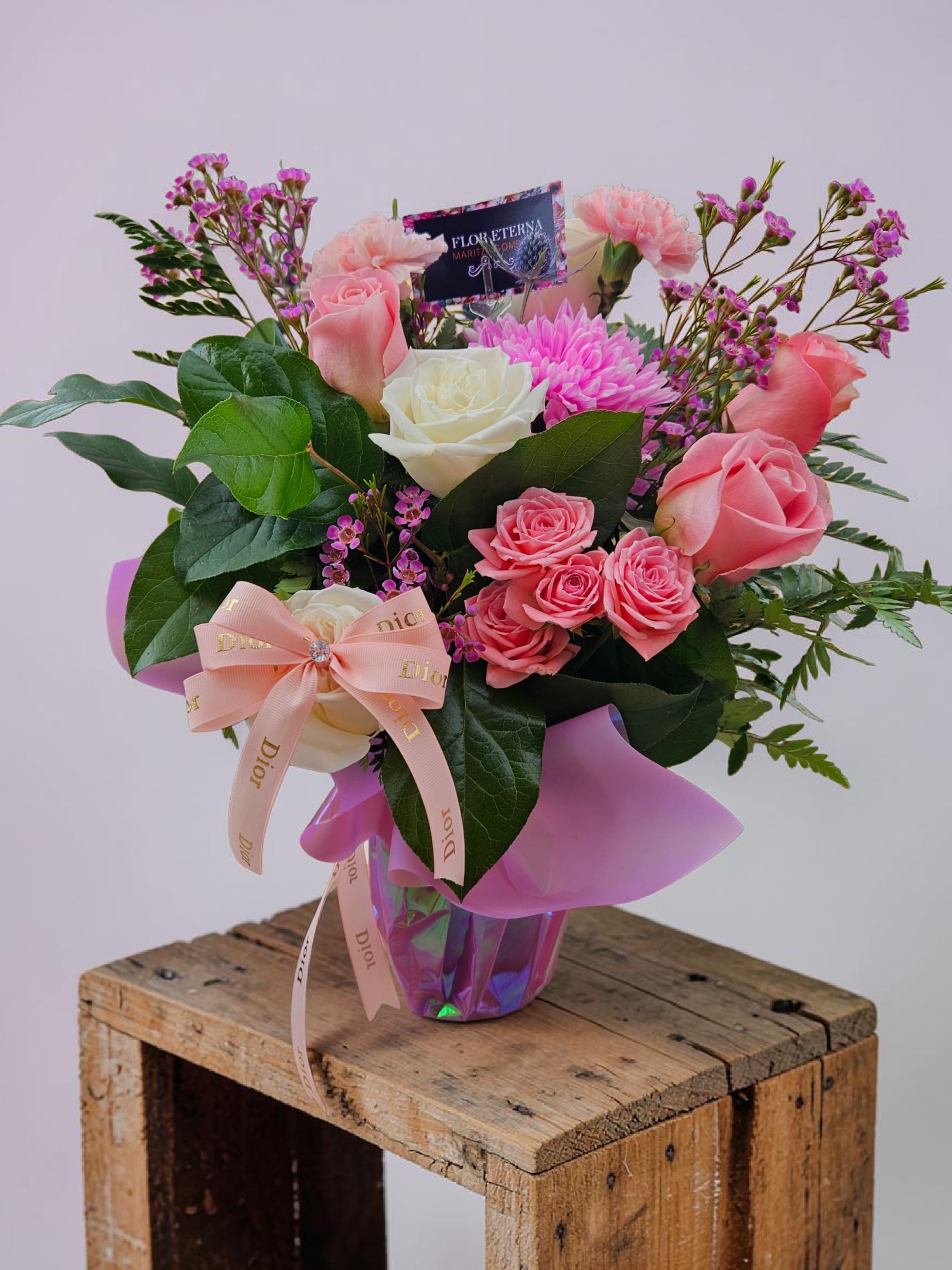 I love You Mom  - Soft pink and white roses take center stage in this "I love You Mom" flower arrangement, making it an ideal Mother's Day bouquet or heartfelt birthday surprise. Designed to make the person you're thinking of feel cherished, the mix of pale pink carnations and a bright pink chrysanthemum-like bloom gives an immediate impression of warmth and celebration-perfect for anniversaries, thank-you moments, or simply saying "I love you."  Layered with small purple filler flowers and large dark green leaves for contrast and structure, the bouquet reads as both lush and polished. It arrives wrapped in pink paper and tied with a pale ribbon bow, then nestles in a faceted iridescent purple glass vase that catches the light like a keepsake. A product tag is included for your message, and the floral wrap keeps the presentation gift-ready on arrival.  This arrangement suits a mom, grandmother, or any woman who delights in soft, romantic color palettes; it conveys affection without being overly formal. Vase included-no extra work needed-so the recipient can display the bouquet immediately and enjoy its layered textures and gentle hues for days.