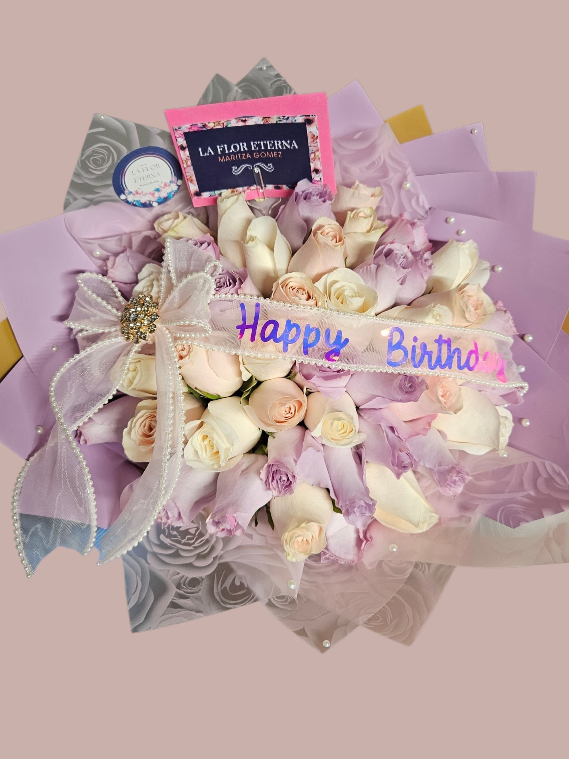 Suspiro Violeta - A rounded birthday bouquet of roses in soft blush, cream and lavender that reads like a pastel daydream - perfect for a birthday, anniversary, or any occasion where you want the person you're thinking of to feel cherished. The dense, hand-tied cluster of roses creates a full, velvety dome that makes the recipient feel seen and celebrated the moment they open it.  Pale pink, blush, cream and lavender roses are wrapped in layered pastel papers (pink, purple and gold-toned panels) and finished with sheer ribbon, satin ribbon accents, pearl trim and an ornamental jeweled bow for a polished, gift-ready presentation. A reflective "Happy Birthday" band and small branded card are tucked into the arrangement; optional gift wrap and a rhinestone brooch add extra sparkle. Size and exact rose count vary by selection - each bouquet arrives carefully prepared to maintain its rounded shape and immediate visual impact.  Ideal for sending warm birthday wishes, marking an anniversary, or saying "I love you" with subtle romance; the soft color palette suits both intimate and celebratory moments. Order with a personalized gift card and choose add-ons like extra decorative ribbon or a brooch to tailor the presentation - roses arrive ready to display in water and will look best when recut and placed in a clean vase upon arrival.