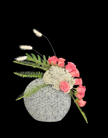 Garden in Balance - Bright pink roses arranged along one side of a rounded ceramic vase create a fresh, romantic focal point-perfect as a birthday bouquet or an anniversary flower arrangement that feels modern and thoughtfully composed. The asymmetrical silhouette and the black geometric pattern on the vase give this gift a contemporary edge, so the person you're thinking of will feel both seen and celebrated.  Soft white filler blooms and three tall, fuzzy stems add airy texture and gentle height, while long green fern fronds reach outward for a loose, garden-fresh look. The ceramic vase provides a sturdy base and stylish contrast with its black detailing; colors here are vivid pink, clean white, leafy green and touches of black for a chic finish. This piece works beautifully as a gift or a home display-no extra styling needed-just trim stems occasionally and refresh the water to keep the arrangement looking its best.