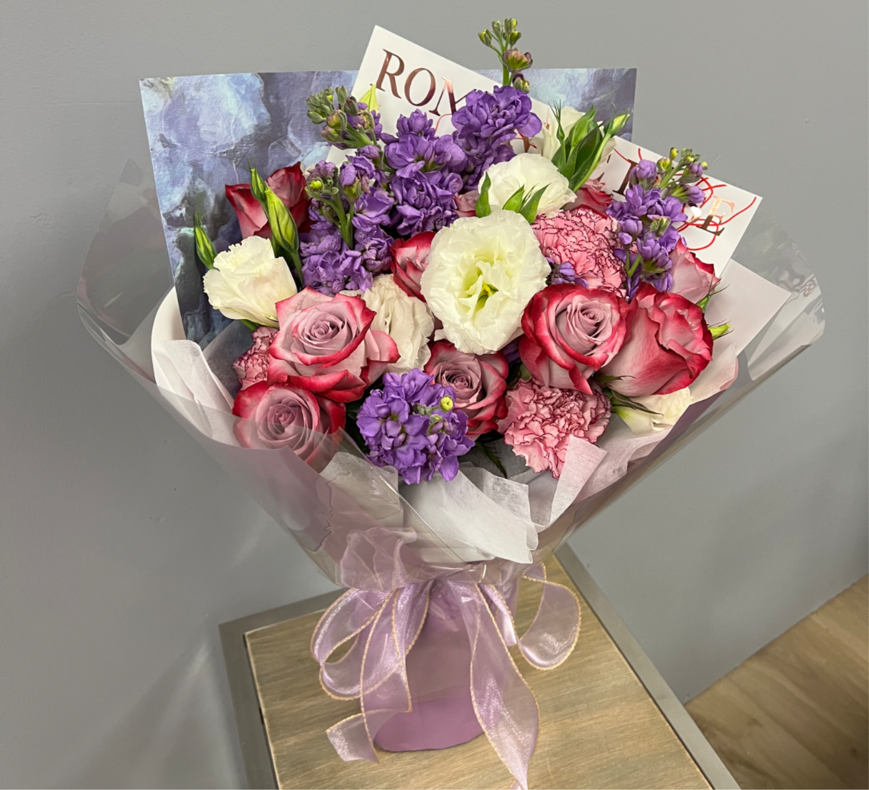 Hand-tied bouquet of pink roses, purple flowers, and white blooms wrapped in pink paper with a sheer ribbon.