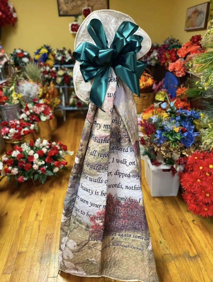 If You Could See Me Now Quilted Throw - A tall, formal sympathy floral tribute crafted to honor a loved one with quiet dignity - perfect as a funeral or memorial arrangement. This standing memorial piece is wrapped in a printed fabric wrap with sentimental text and finished at the top with a large teal satin bow, giving it a respectful, commemorative presence that reads as a heartfelt expression of condolence.