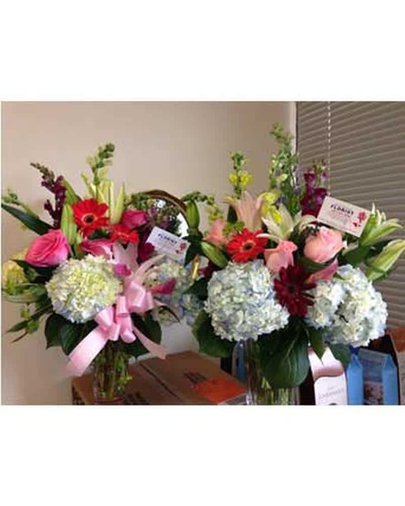 Two tall mixed flower arrangements with hydrangeas, pink roses, lilies, and red gerbera daisies in glass vases.