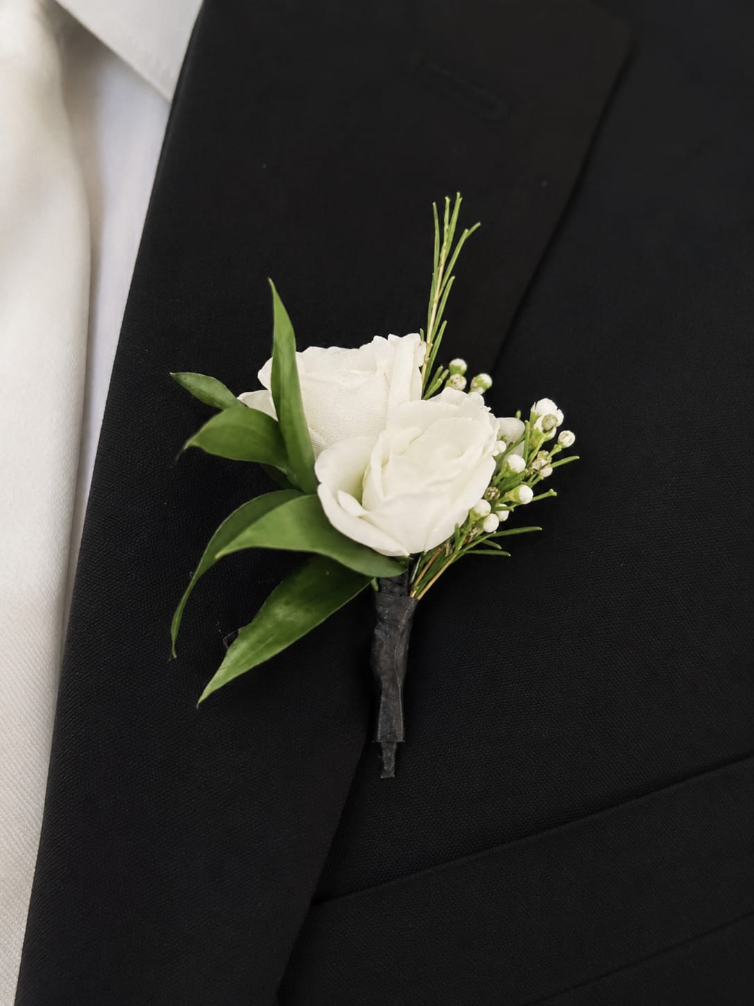 Ivory Whisper Boutonniere - The Ivory Whisper Boutonniere is a sophisticated floral accessory designed for the groom who values understated elegance and refined details. At its center is a fresh white rose, accented with delicate waxflower and carefully selected Italian ruscus foliage.  The soft ivory and fresh green palette creates a timeless look that complements classic, garden-style, romantic, and modern weddings alike. Its natural textures and airy design bring a touch of sophistication while remaining effortlessly elegant.  Handcrafted with care, each boutonniere is designed to coordinate beautifully with the Ivory Whisper Bridal Bouquet, creating a harmonious floral story for the couple’s wedding day. Perfect for the groom, groomsmen, fathers, and other members of the wedding party.