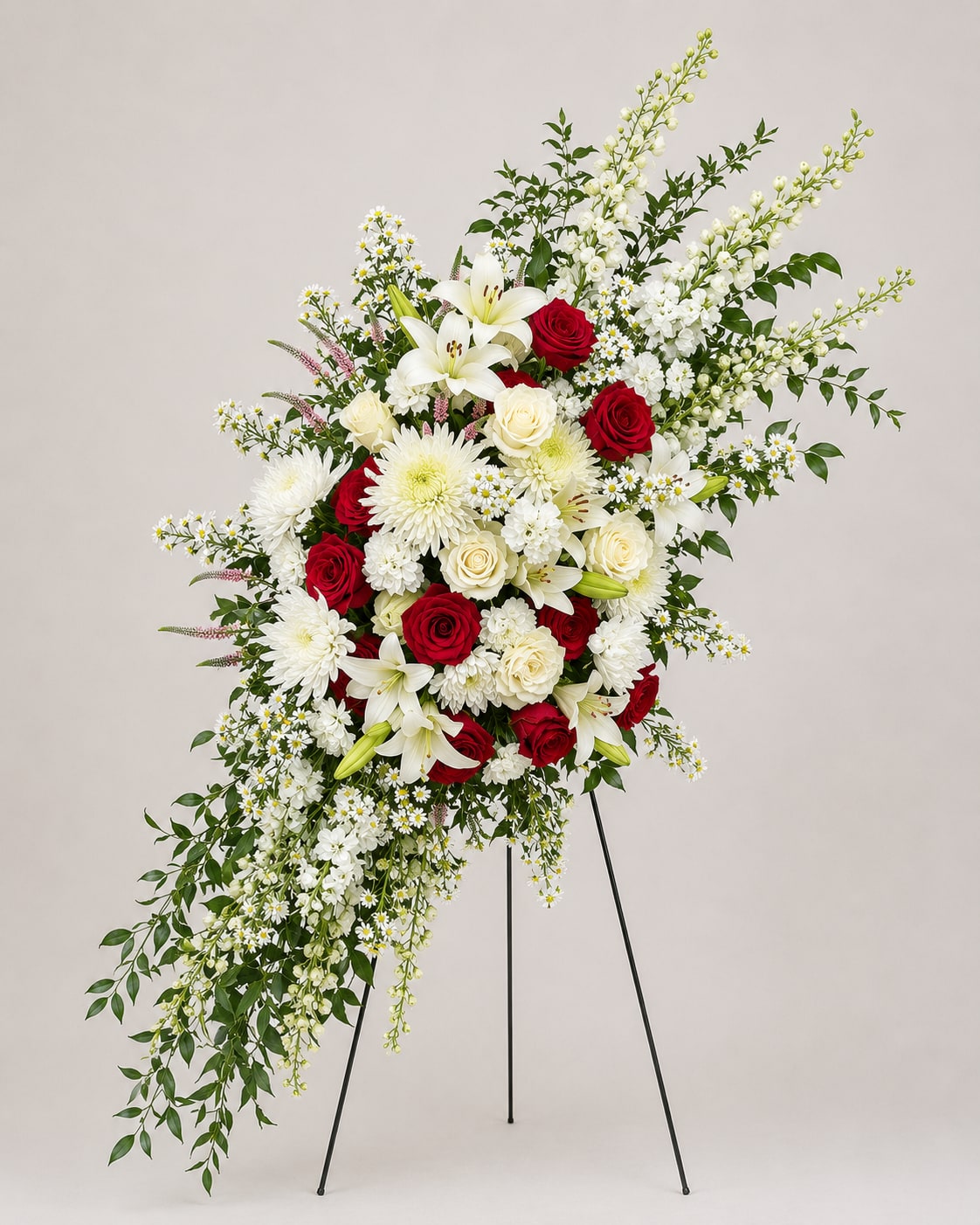 Eternal Grace Tribute Spray - The Eternal Grace Tribute Spray is a sophisticated and deeply meaningful arrangement created to honor life with elegance and respect. This design features a curated selection of premium white lilies, roses, chrysanthemums, and delicate seasonal blooms, complemented by rich red roses symbolizing enduring love and remembrance. The composition is styled in a soft, natural flow with lush greenery, creating movement, depth, and a refined visual presence. Presented on a wooden easel stand, this standing spray offers a graceful and elevated display, making it a perfect choice for funeral services, memorial ceremonies, or graveside tributes. Each piece is thoughtfully handcrafted to ensure a unique and polished result, reflecting both beauty and sincerity in every detail.