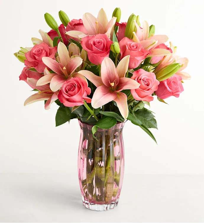 Blush Elegance Bouquet  - Bright pink roses mingle with pale pink-and-cream lilies in a soft, romantic silhouette-an ideal birthday bouquet, anniversary flowers, or Mother's Day arrangement that says "I'm thinking of you" with effortless charm. The layered look, with star-shaped lilies lifting above open roses, creates a lively contrast of textures that will make the recipient feel genuinely celebrated and seen.  Hand-gathered into a clear pink glass vase, the arrangement reads fresh and feminine without feeling fussy; the vase's subtle blush tint complements the blooms rather than competing with them. Expect a full, rounded shape roughly the size shown in the photos, with lush greenery tucked among the stems to enhance color and depth. This versatile everyday gift arrives ready to display-simple care like fresh water and a cool spot will keep the roses and lilies looking lovely for days.