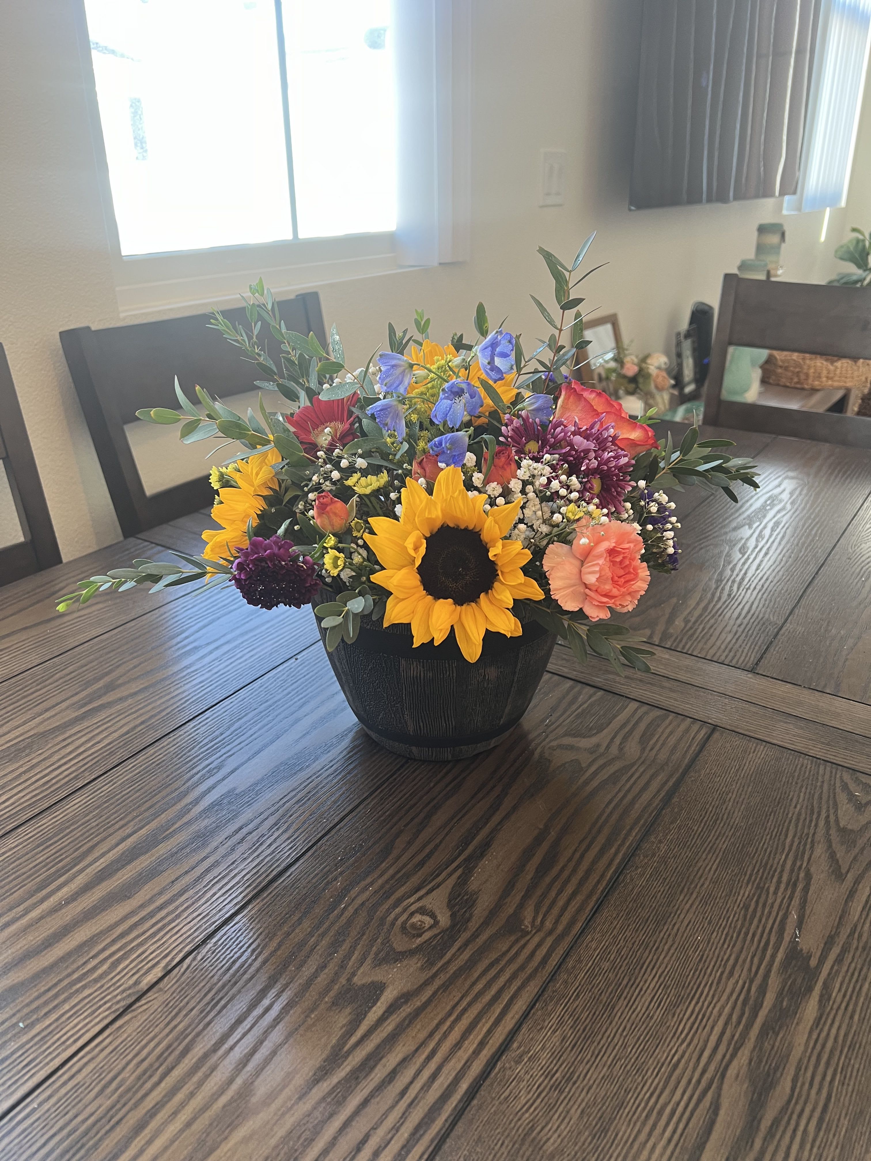 Spring Farm collection - Golden sunflowers mingle with purple and burgundy chrysanthemums and soft pink carnations in a lively Spring Farm collection arrangement-perfect as a birthday bouquet, a Mother's Day surprise, or a "just because" pick-me-up. The bright yellow, pink, blue-tinged accents and white filler flowers create a cheerful, rustic look that makes the recipient feel genuinely celebrated and seen.  Arranged low and full in a matte black vase, the composition spreads outward with airy greenery for a casually abundant centerpiece. Textures range from the bold sunflower faces to the ruffled carnations and spidery mums, with delicate white fillers adding lightness; the palette reads as mixed color with strong seasonal cheer. Ideal for spring and summer tables, thank-you gifts, or retirement celebrations, this arrangement arrives ready to display-no extra styling required-and will keep its look with regular fresh water changes.