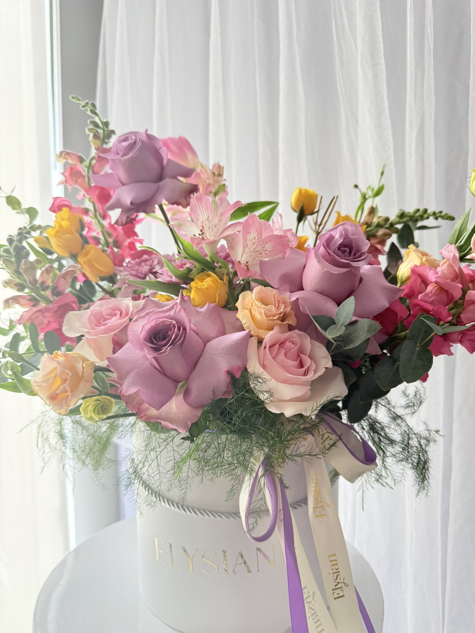 Monet’s Garden - A joyful blend of color, texture, and movement — this garden-inspired arrangement brings together premium roses in mauve and blush tones with vibrant seasonal blooms in soft pinks, peaches, and sunny yellows. Designed in our signature keepsake box, it feels abundant, artistic, and effortlessly elegant.  Layered with delicate florals and fresh greenery, this piece captures the beauty of a blooming garden while offering the modern convenience of a floral box — no vase needed. Perfect for celebrations, heartfelt gestures, birthdays, or brightening someone’s day with something truly special.  Each arrangement is thoughtfully designed to feel organic and luxurious, with flowers placed to create natural flow and dimension from every angle.  What’s included: • Premium mauve and blush roses • Seasonal mixed blooms and textural accents • Fresh greenery for movement and depth • Signature keepsake floral box with ribbon • Designed in floral foam for easy care (no rearranging required)  Disclaimer: Flower varieties and shades may vary slightly depending on seasonal availability while maintaining the same color palette, style, and overall fullness.