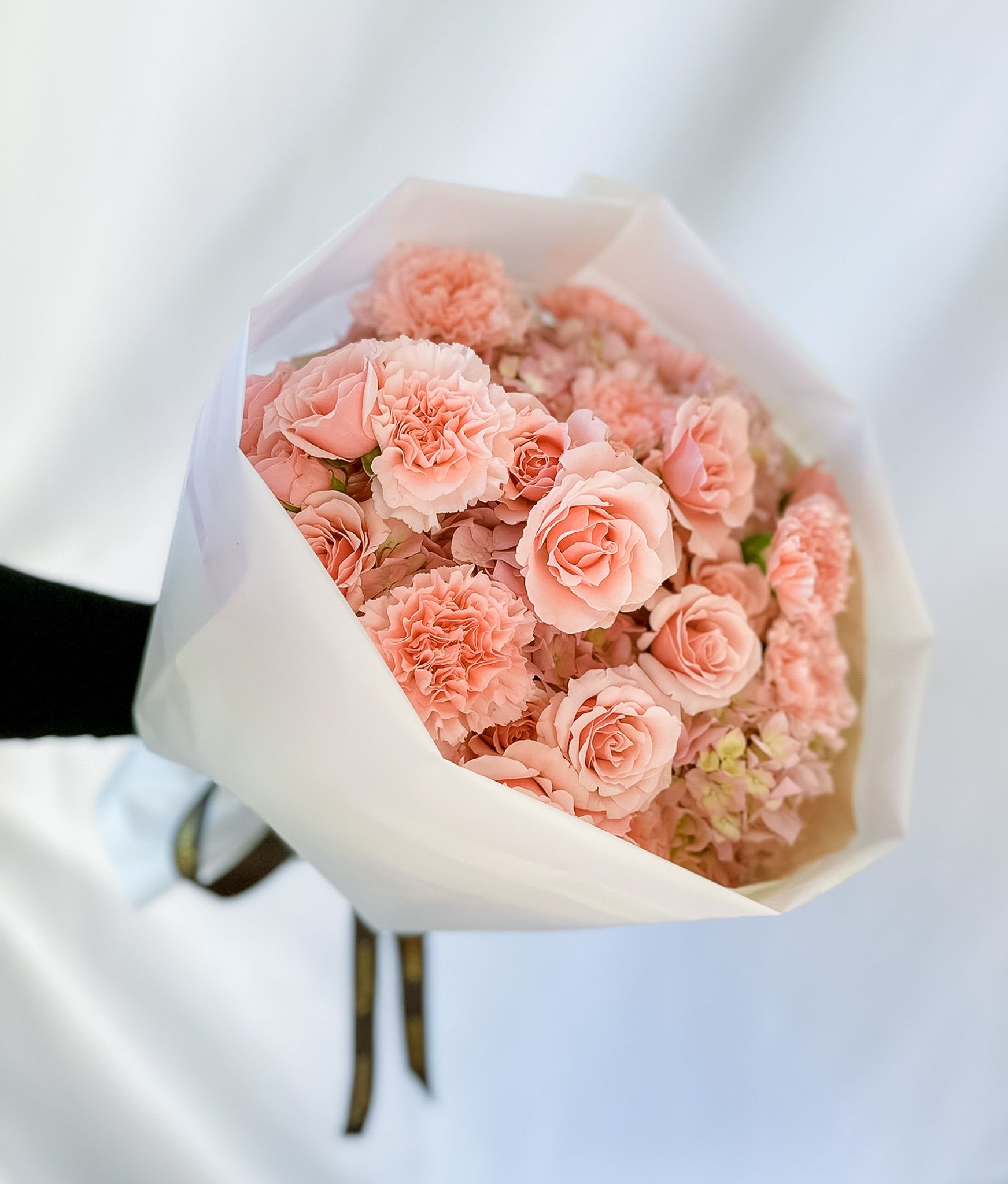 Pretty in Pink - Our best-selling creation is designed to capture hearts. This round arrangement showcases the soft, cloud-like beauty of pink hydrangeas, the delicate allure of spray roses, and the textured sweetness of carnations—all in harmonious shades of pink.  Wrapped in luxury paper and tied with a satin ribbon, this bouquet exudes romance and sophistication. Perfect for Valentine’s Day, anniversaries, or simply as a heartfelt gesture, it’s a gift that speaks of love and thoughtfulness in every petal.  Let this exquisite arrangement bring joy, elegance, and whimsy to someone’s day.  Disclaimer  Flower varieties and exact shades may vary based on seasonal availability. Substitutions may be made while maintaining the overall color palette, style, and value of the arrangement to ensure the freshest blooms and the highest design quality.