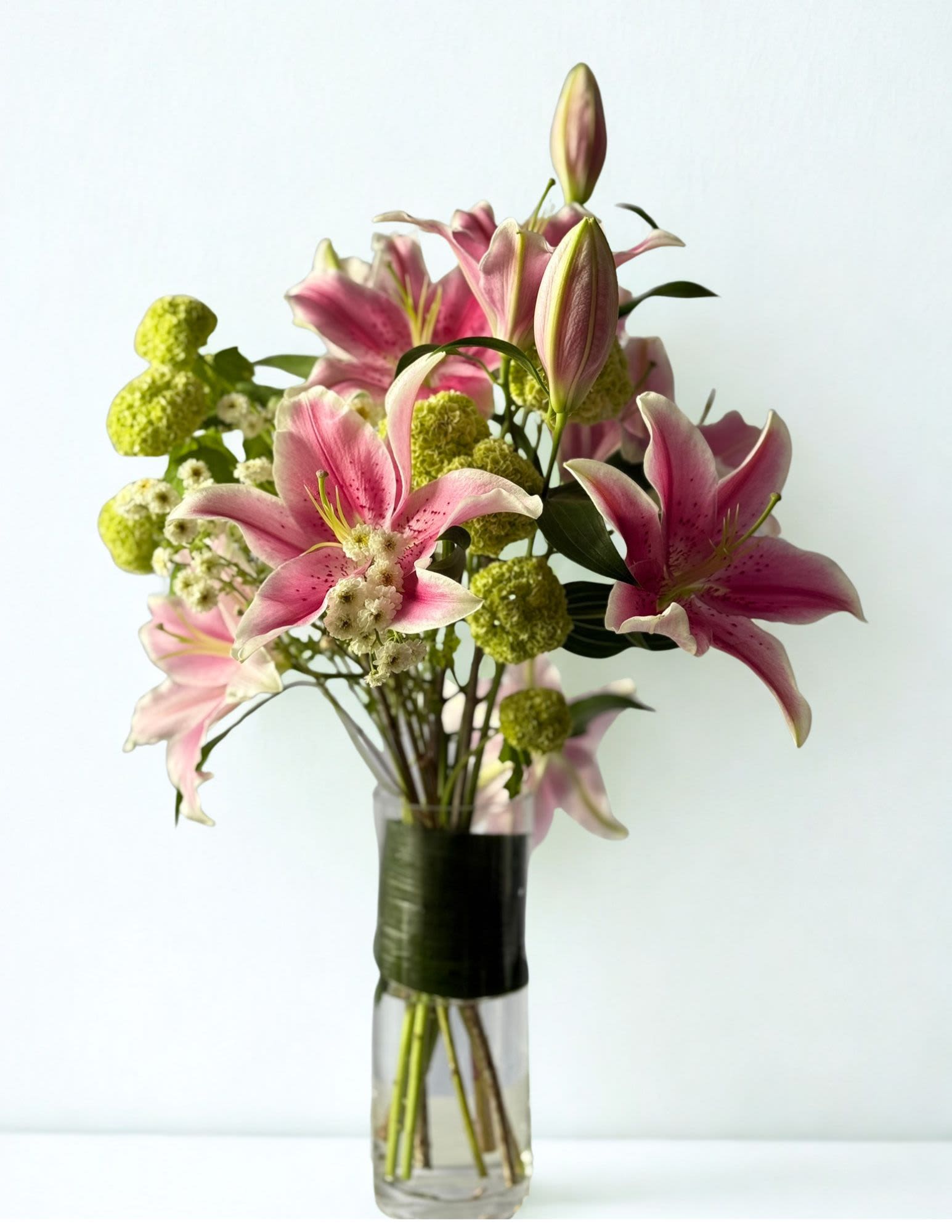 Oriental Lillies - An elegant vase arrangement featuring fragrant pink oriental lilies paired with playful green viburnum balls and delicate white accent blooms. Designed with a modern airy style, this arrangement brings together soft romance and fresh garden charm — perfect for birthdays, celebrations, thank you gifts, or simply brightening someone’s day. As the lilies bloom open, the arrangement transforms beautifully over the following days, creating a stunning statement in any space.  Some lilies may arrive in a more closed stage at the time of delivery. This is intentional to ensure maximum freshness and longevity. Over the next couple of days, the lilies will gradually bloom open beautifully, allowing you to enjoy the full transformation of the arrangement.