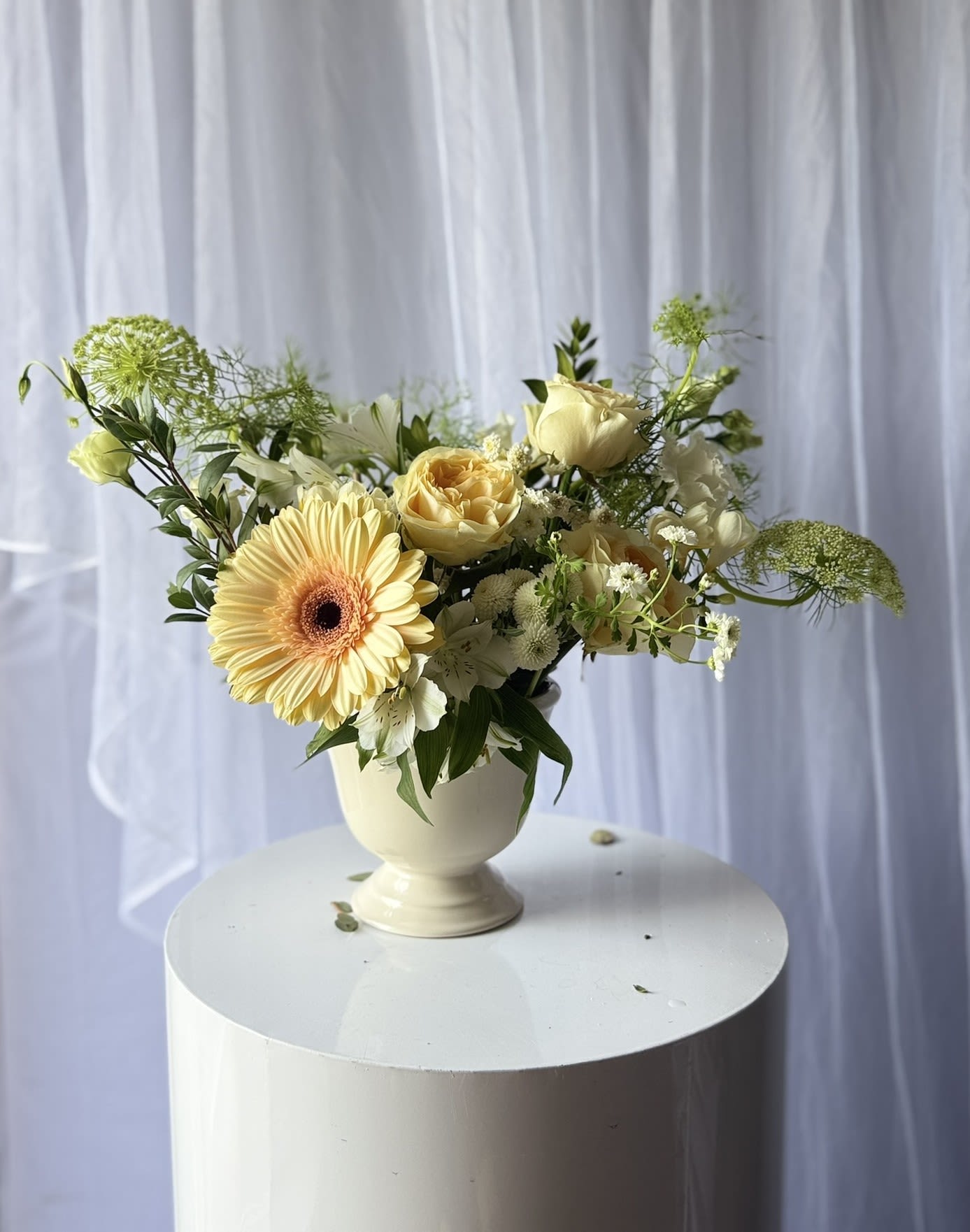 Buttercream bloom - A charming garden-style arrangement featuring soft buttercream roses, a cheerful pastel gerbera daisy, delicate white blooms, and airy seasonal greenery arranged in an elegant ceramic vase. Designed with a whimsical, freshly gathered feel, this piece brings warmth, light, and effortless beauty to any space.  Perfect for birthdays, thank you gifts, new baby celebrations, or simply brightening someone’s day, this arrangement blends soft neutral tones with natural movement for a timeless and uplifting design.