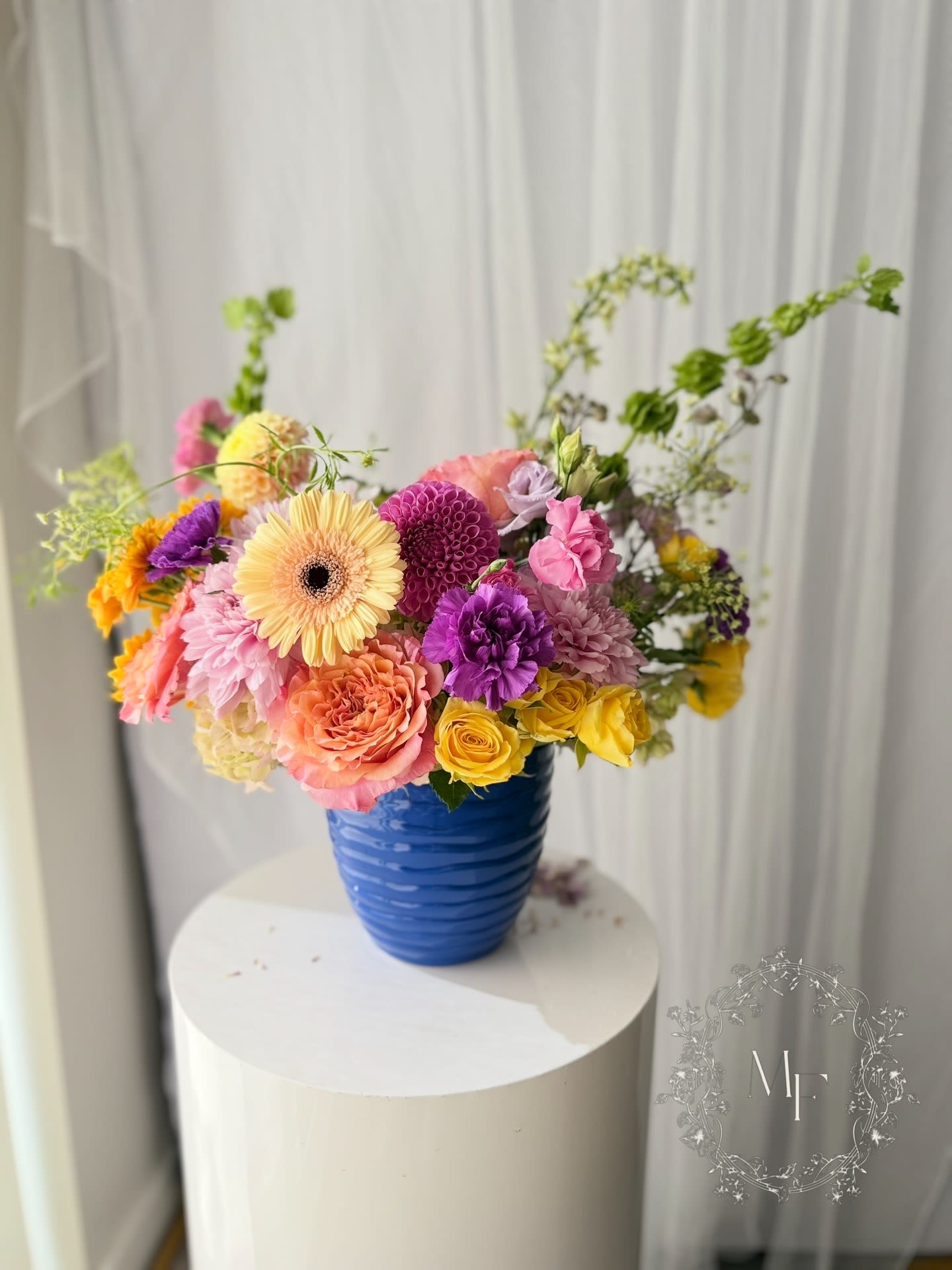 La Fête - Housed in a striking cobalt blue ceramic vase, this arrangement brings together peachy garden roses, sunshine yellow spray roses, magenta ball dahlias, peach gerberas, vivid purple carnations, blush lisianthus, orange gerberas, and wild trailing hops vine — each stem chosen to dance alongside the next, never to compete.