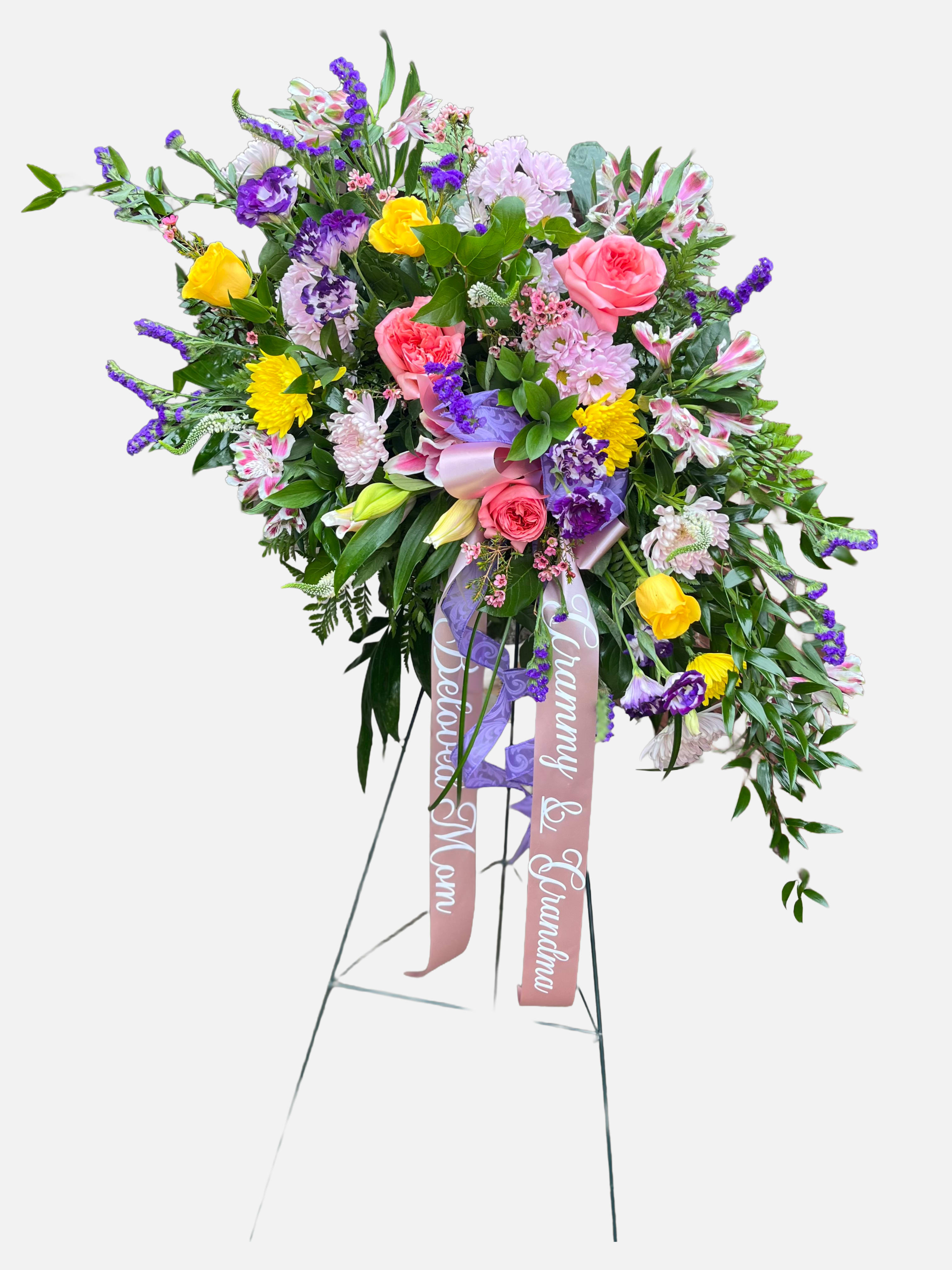 Beautiful Grace - A tall, uplifting standing spray designed for a funeral or memorial service, Beautiful Grace brings a gentle, comforting presence to a farewell. The vertical silhouette on a metal easel makes this sympathy arrangement easy to display at a service or graveside, and the bright pinks, soft pastels and cool purples together create a hopeful, consoling palette that will help the person you're honoring feel remembered and cherished.  This arrangement is densely layered with pink roses, purple lisianthus, yellow chrysanthemums and alstroemeria, plus stock and lilies for soft texture and subtle fragrance. Flowers cascade in a full, graceful shape with a large multi-loop lavender-and-pink bow at the center and two printed memorial ribbons trailing down the front, giving the piece a formal, finished look appropriate for commemorations.  Perfect for expressing sympathy, celebrating a life, or sending condolences to the family, Beautiful Grace reads as both respectful and gently joyful-an arrangement that comforts while celebrating fond memories. Mounted on a sturdy metal easel for clear visibility at the service; please let us know any personalization requests for the printed ribbons when ordering.