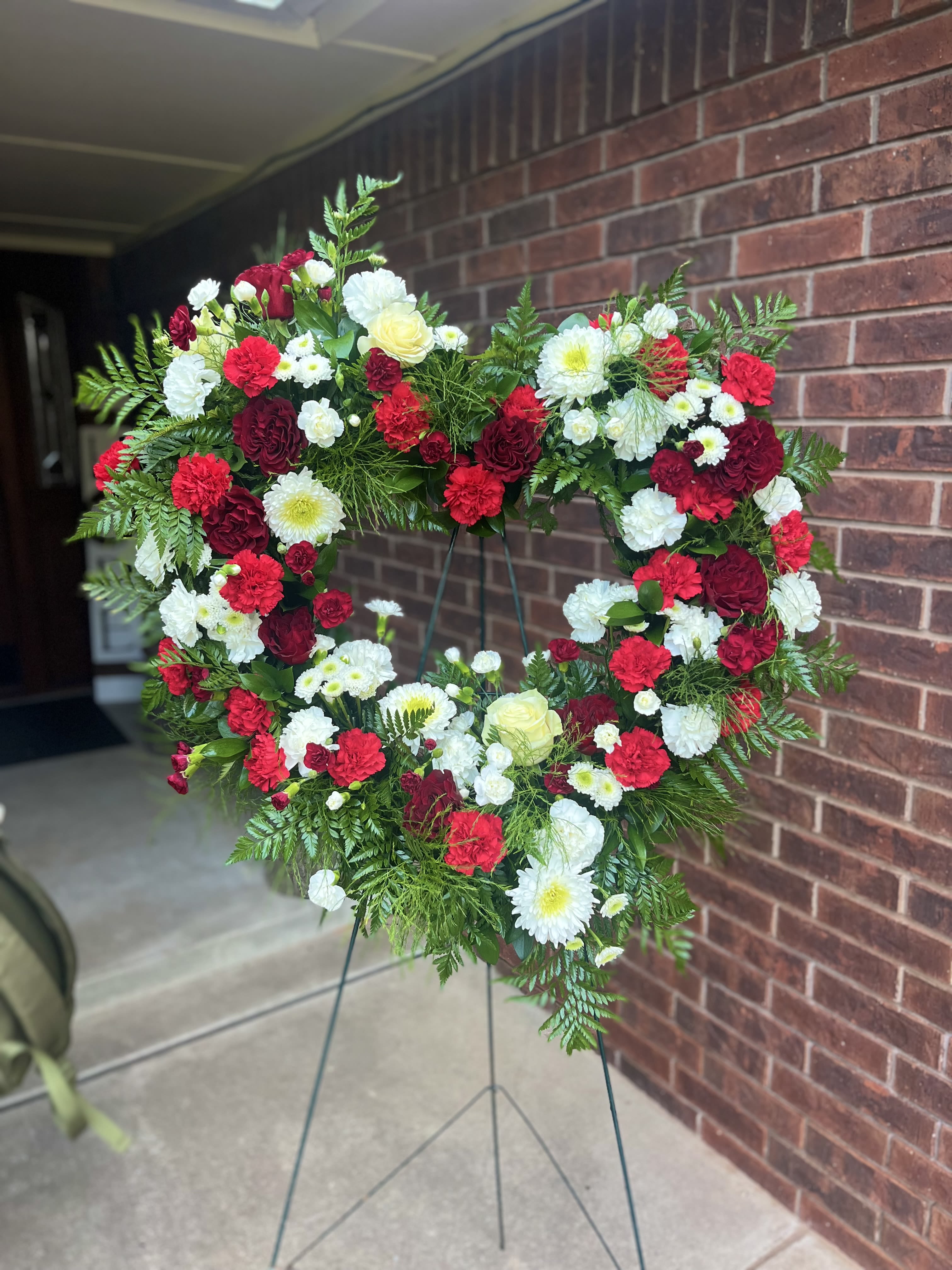 18” Red and White Heart tribute - A heart-shaped standing wreath in rich reds and crisp whites designed for sympathy services and memorials - the perfect way to honor a loved one at a funeral or graveside tribute. The dominant red and white palette reads as reverent and heartfelt, while the touch of soft yellow brings a gentle note of warmth that helps the arrangement feel consoling rather than stark.  This large wreath is built on a sturdy metal easel and shaped with abundant greenery that outlines the heart and gives the piece a full, textured silhouette. Clusters of roses, carnations, chrysanthemums, and daisies are interwoven across the form so the mix of petals and foliage reads cohesive from every angle; red blooms provide dramatic depth, white flowers offer calm clarity, and yellow accents add subtle contrast.  Made to make the person you're remembering feel honored, this standing heart wreath is intended for formal services, memorial displays, or placement at a graveside. The metal easel provides stable support for display; please let us know service delivery details when ordering so we can coordinate placement and timing.