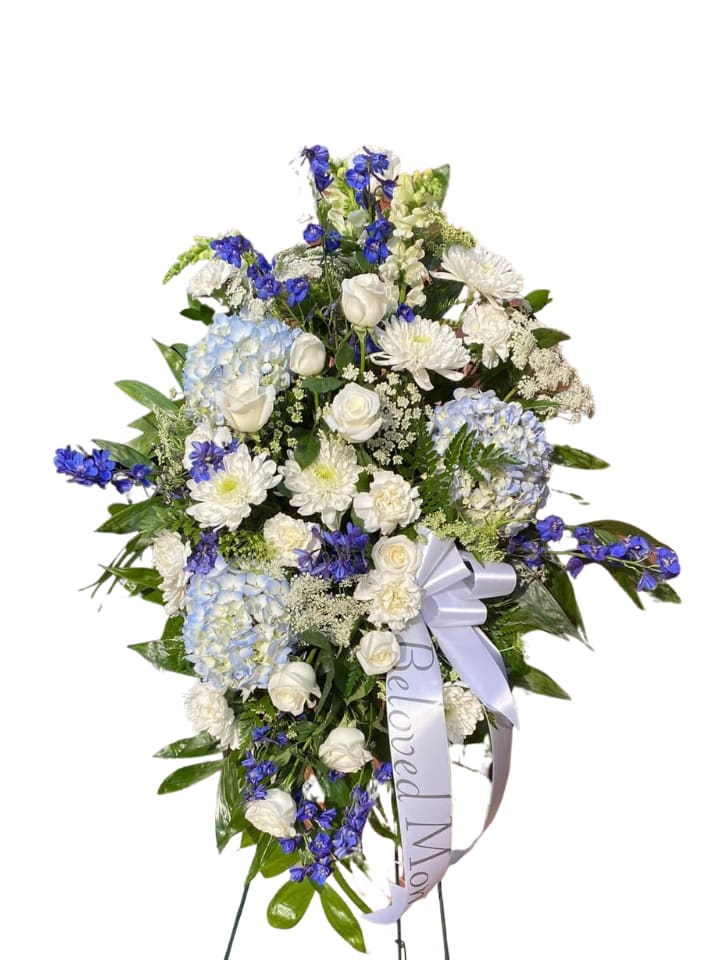Tranquil waters standing spray - Soft blue hydrangeas and bright delphinium create a calming wash of color in this funeral standing spray, a serene tribute perfect for a memorial or funeral service honoring a beloved mother. The oval-shaped arrangement reads as peaceful and respectful from a distance, the white roses and chrysanthemums bringing classic dignity while the blue accents introduce a gentle, soothing presence.  Close-up, the spray combines white roses, chrysanthemums, and carnations with soft blue hydrangeas, bright blue delphinium, and delicate filler blooms for layered texture and depth. It comes mounted on a metal easel and finished with a white satin bow plus a white memorial ribbon printed with a commemorative message, giving the piece a formal, finished look that carries the sentiment clearly.  Designed to be placed beside the casket or at the service entrance, this arrangement comforts those who see it and offers a visible expression of love for the person you're honoring. Available in a tall, oval silhouette suitable for viewing from standing or seated positions; please contact us if you need delivery or sizing details for the service location.