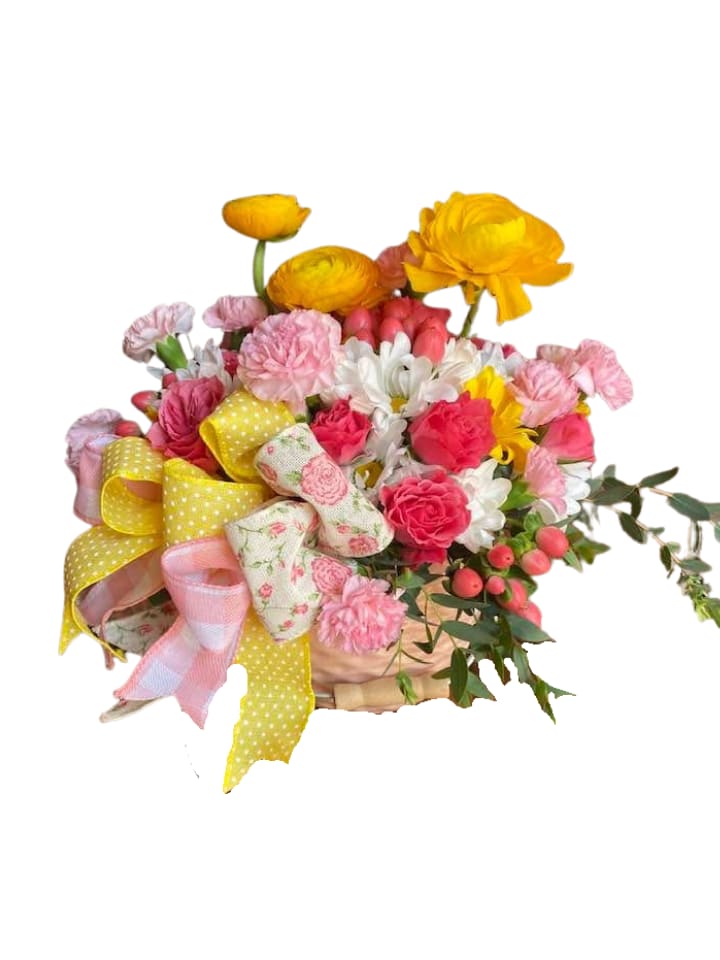  Rasberry Lemonade - Bright clusters of bright pink carnations and roses give this spring basket an instant joyful burst-the perfect birthday or welcome baby bouquet. Designed as a compact, low arrangement in a light woven basket, it reads like a walk through a sunny cottage garden and is ideal for celebrations, sending encouragement, or brightening someone's desk or kitchen table.  The mix pairs hot pink carnations and roses with golden yellow ranunculus, crisp white daisies, and small coral berry accents for contrast. Large decorative fabric ribbons-yellow polka dot, pastel plaid, and a floral print-are tied at the front to add playful texture and a handcrafted feel. Because the flowers are arranged low and full, the piece delivers abundant color without towering stems, making it easy to display in small spaces.  Give this arrangement to someone who deserves a bright moment-a friend celebrating a milestone, a neighbor in need of cheer, or welcome a new baby. The woven basket and sturdy ribbons arrive ready to display; for best enjoyment, keep blooms in a cool spot away from direct sun and refresh water as needed.