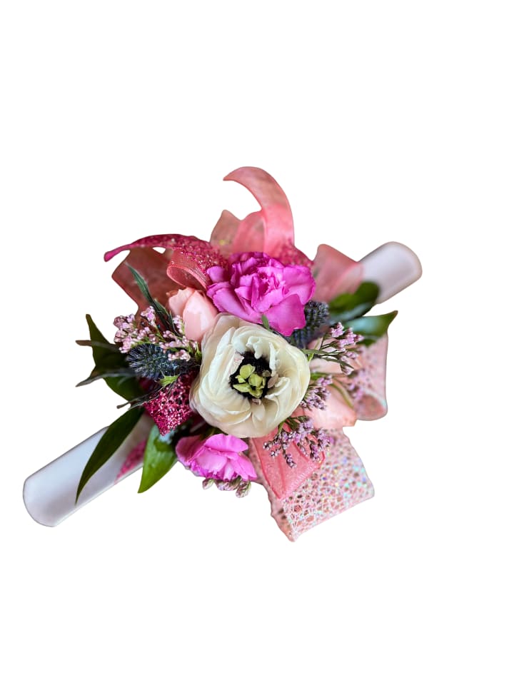 Hope Floats - A creamy white ranunculus takes center stage in this compact wrist corsage, surrounded by vibrant pink blooms. The tightly clustered flowers create a lively burst of pink, white, and lavender, making the recipient feel noticed and celebrated.  The arrangement is presented with a wide, glittery pink ribbon that forms a shimmering backdrop. It’s attached to a rolled white paper scroll and paired with a small, iridescent patterned gift box for an extra-special reveal. This petite and polished piece is designed for easy gifting. Its compact size makes it ideal for desk or bedside display, and the ribbon and box ensure it arrives ready to give.  Choose this arrangement when you want a playful, gift-ready bouquet to mark congratulations, a milestone, or a cheerful “thinking of you.” No special care is required beyond keeping the blooms cool and out of direct sunlight. The compact format makes it effortless to enjoy right away.