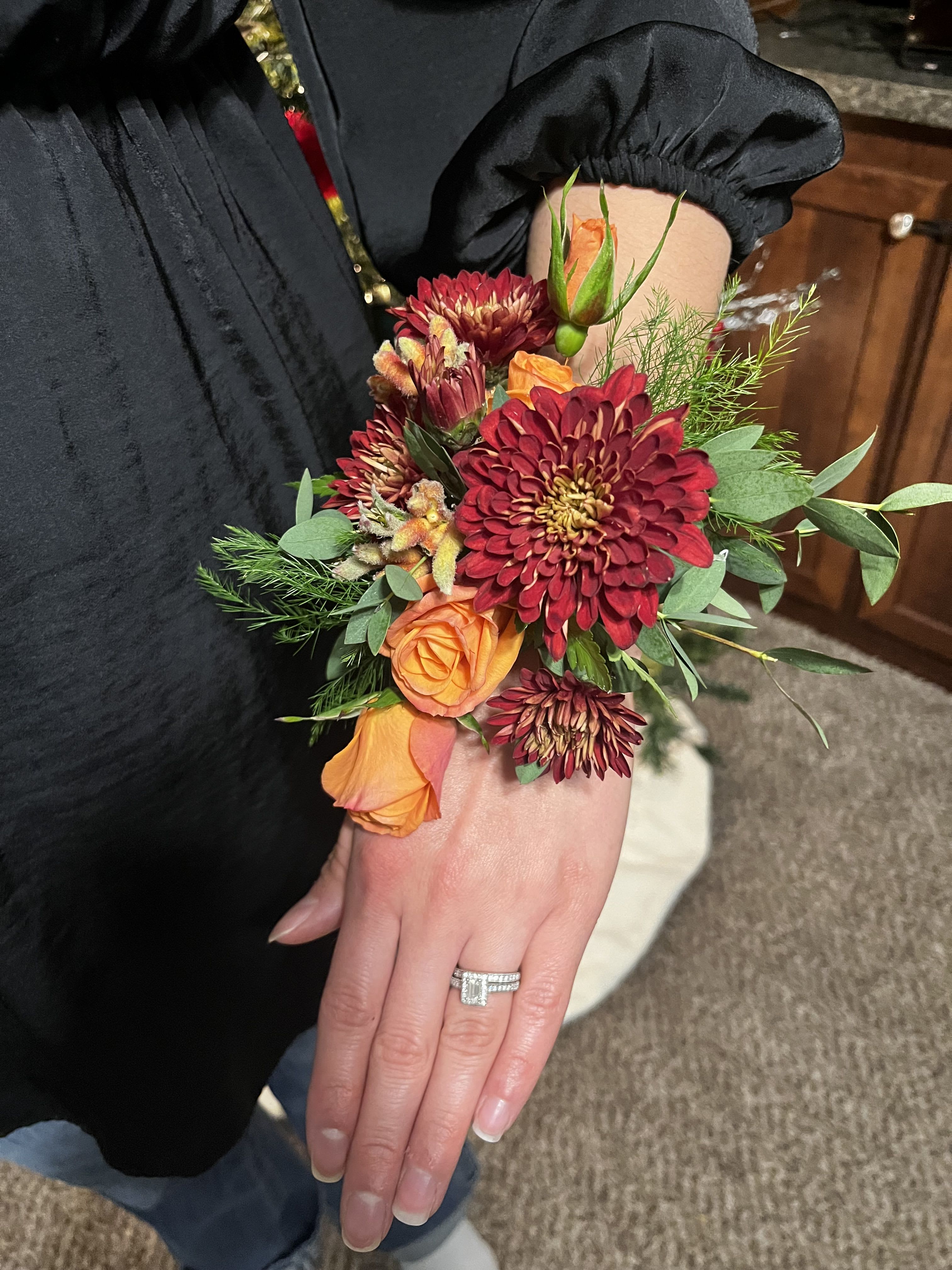 Elite Corsage - Deep burgundy chrysanthemums and bright orange roses make this wrist corsage a striking choice for prom, a fall wedding, or any autumn celebration where the recipient should feel noticed. The warm, festive color story-rich red and orange set off by green foliage-reads elegant against a dark dress and sends the message that the person you're honoring is the evening's focal point.  Arranged tightly along a comfortable wrist corsage band, the flowers sit close for a polished, wearable silhouette. Feathery and broad green foliage adds rustic texture and fullness without overpowering the blooms; the overall scale is compact and designed to move with the wearer. This is the kind of corsage that complements formal attire and makes her feel genuinely celebrated-order with confidence for proms, bridesmaids, or special autumn events.