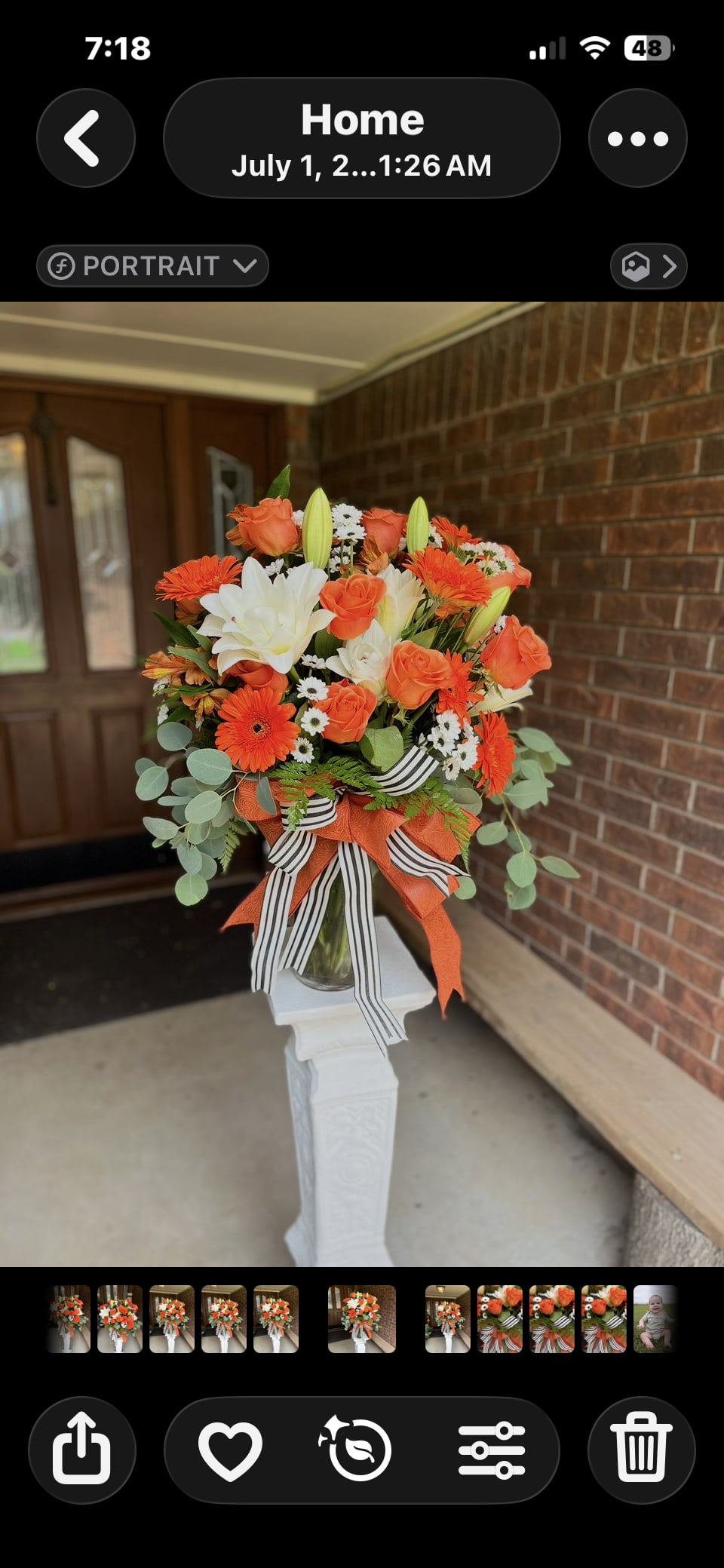 Collegiate Splendor - Bright orange roses and gerbera daisies meet large white lilies and cheerful white daisies in a tall celebratory bouquet designed as an entrance-piece for special moments. This flower arrangement and centerpiece works beautifully as a birthday bouquet, autumn event display, or graduation/ceremony arrangement-the bold orange and white color story reads energetic and festive, so the person you're honoring will feel noticed and celebrated.  The flowers are gathered densely into a rounded, full top sitting atop a clear glass vase, creating strong visual impact and height. Orange roses and gerbera daisies provide warm, textured focal blooms, while the white lilies and small white daisies with yellow centers offer contrast and lightness. A layered bow of vivid orange ribbon and a black-and-white striped ribbon wraps the vase neck for a graphic, collegiate-inspired accent; the whole vase is shown on a white decorative pedestal stand, giving it formal presence for entryways, reception tables, or stage décor.  This arrangement is tailored to make a statement at events-think autumn gatherings, festive parties, or graduation ceremonies-where the recipient or honoree should feel genuinely celebrated. The tall vase and pedestal presentation make it easy to place as a focal display; for best longevity keep flowers in fresh water, trim stems at delivery, and place away from direct heat or drafts.
