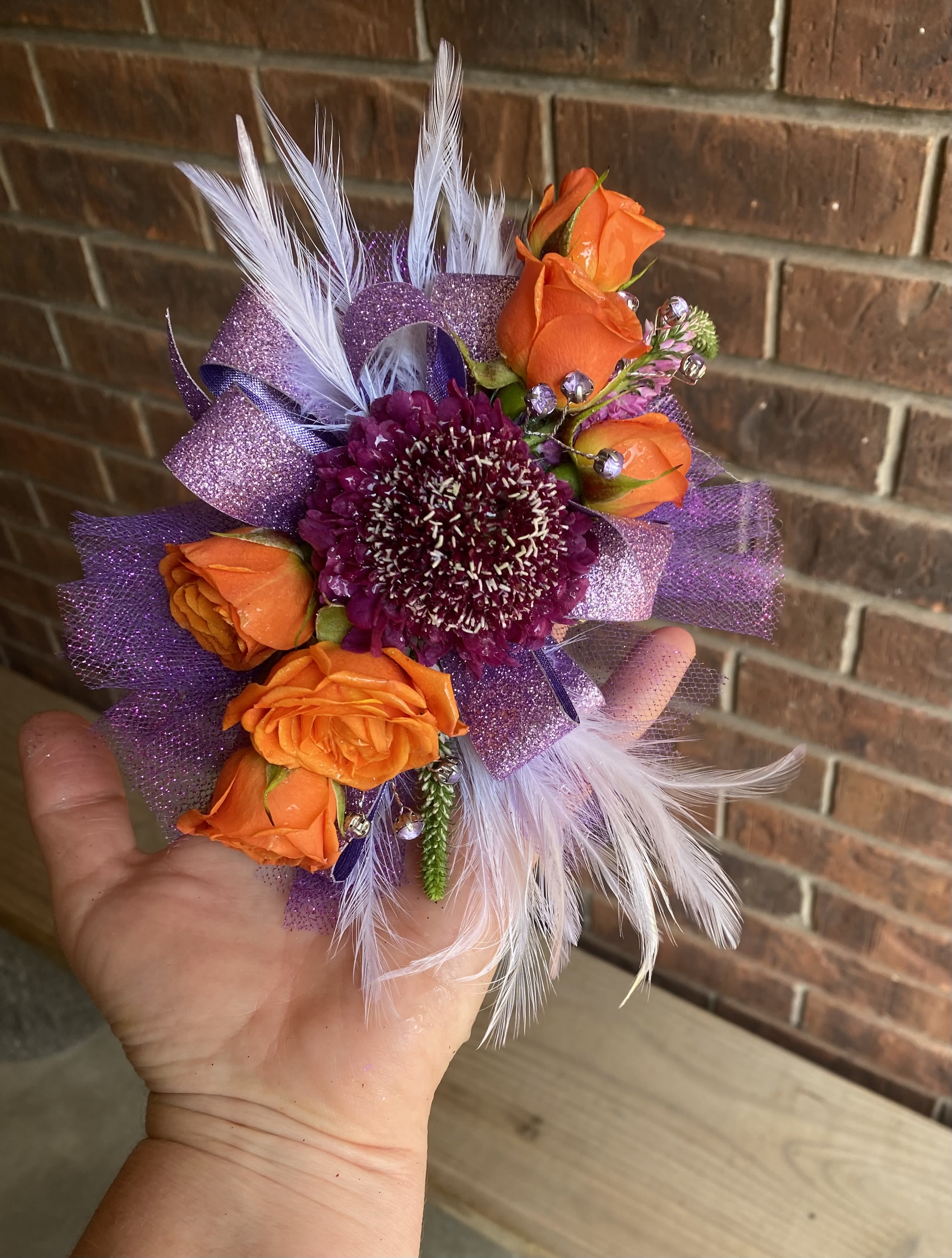 Fancy Corsage - A compact hand-held corsage that pops with color-this fancy corsage pairs orange roses with a single deep purple bloom for a bold, youthful statement perfect for prom, a birthday, or any cheerful celebration. The lavender-purple wrapping and bright contrasts make the recipient feel noticed and celebrated, while the playful mix of textures keeps the look festive rather than formal.  Crafted with orange roses and a central deep purple flower, the arrangement is accented with white feathers, decorative beads, and purple glitter ribbon for sparkle and movement. The small, hand-held scale is designed to sit comfortably in the hand or be carried on the wrist; the lavender-purple wrap ties the palette together and highlights the vivid orange and white accents.  Ideal for the person who loves color and fun-give this as a prom corsage, lively birthday gift, or a celebratory token that reads as joyful and whimsical. Because of its compact size and decorative trims, handle gently and store in a cool place until the moment you give it.