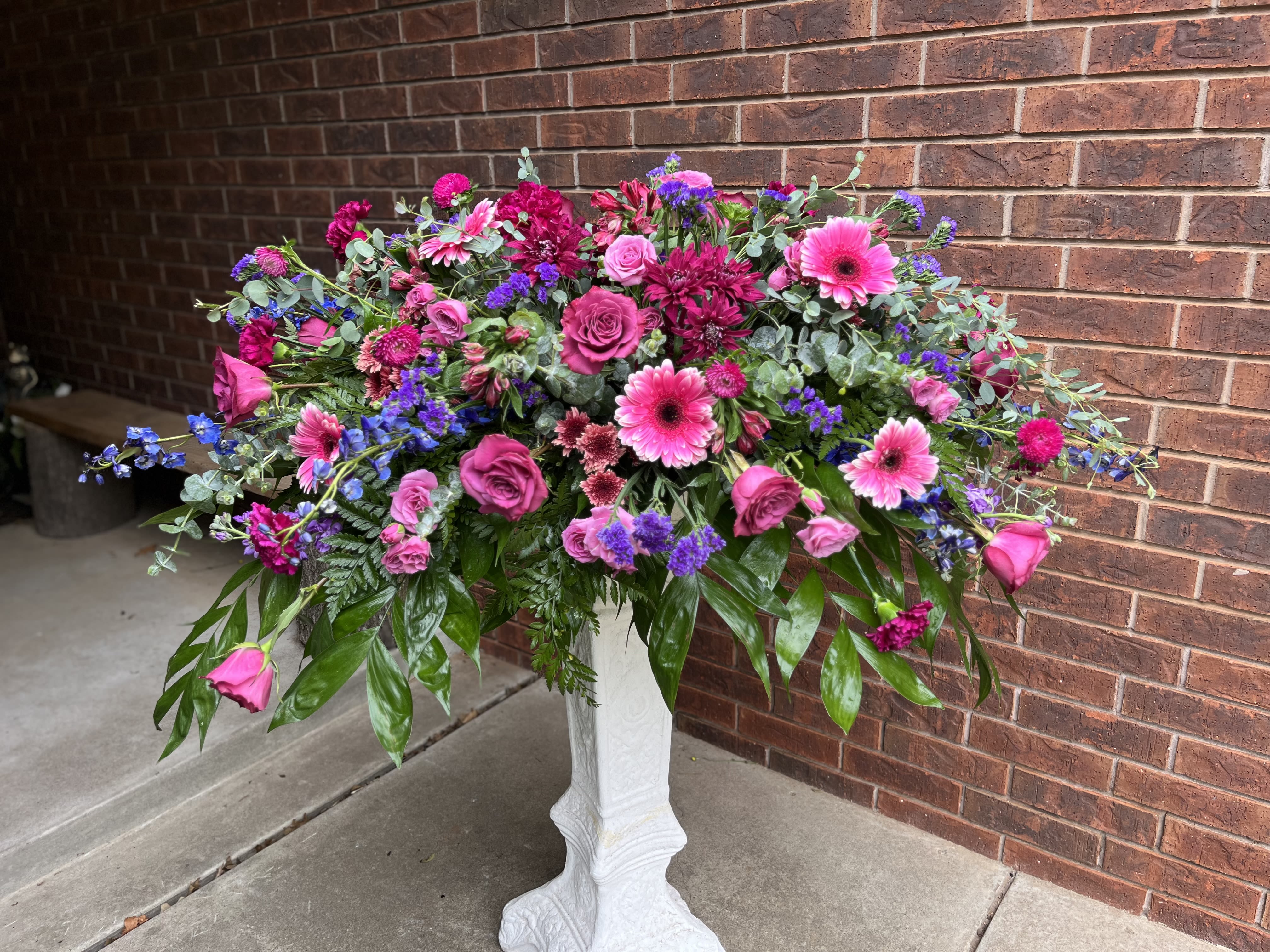 Majesty Tribute - A lush, overflowing tribute arranged on a white pedestal vase, this floral arrangement is a striking choice for a sympathy or memorial setting and also works beautifully for weddings, anniversaries, or milestone birthdays. The Majesty Tribute offers a celebratory yet respectfully elegant presence-its tall, formal silhouette and abundant pink, purple, white, and green palette make the recipient feel honored and remembered.  This large design is densely built with roses, gerbera daisies, carnations, alstroemeria and dianthus, plus delicate filler blooms and broad glossy foliage that cascades over the pedestal for a layered, full look. The mix of soft pinks and purples with eucalyptus accents creates visual contrast while the varied textures-from velvety roses to bold daisy faces-give the arrangement depth and movement. The arrangement arrives in a classic white pedestal vase that elevates the display and makes it suitable for entryways, altars, or reception tables.  Perfect for marking a meaningful moment-whether to commemorate a life, celebrate an anniversary, or add refined floral impact to a wedding-this piece sends a message of warmth and reverence to the person you're honoring. Because of its scale and formal presentation, allow for a prominent spot when you place it; routine care is simple-fresh water and a cool location will help blooms remain vibrant for as long as possible.