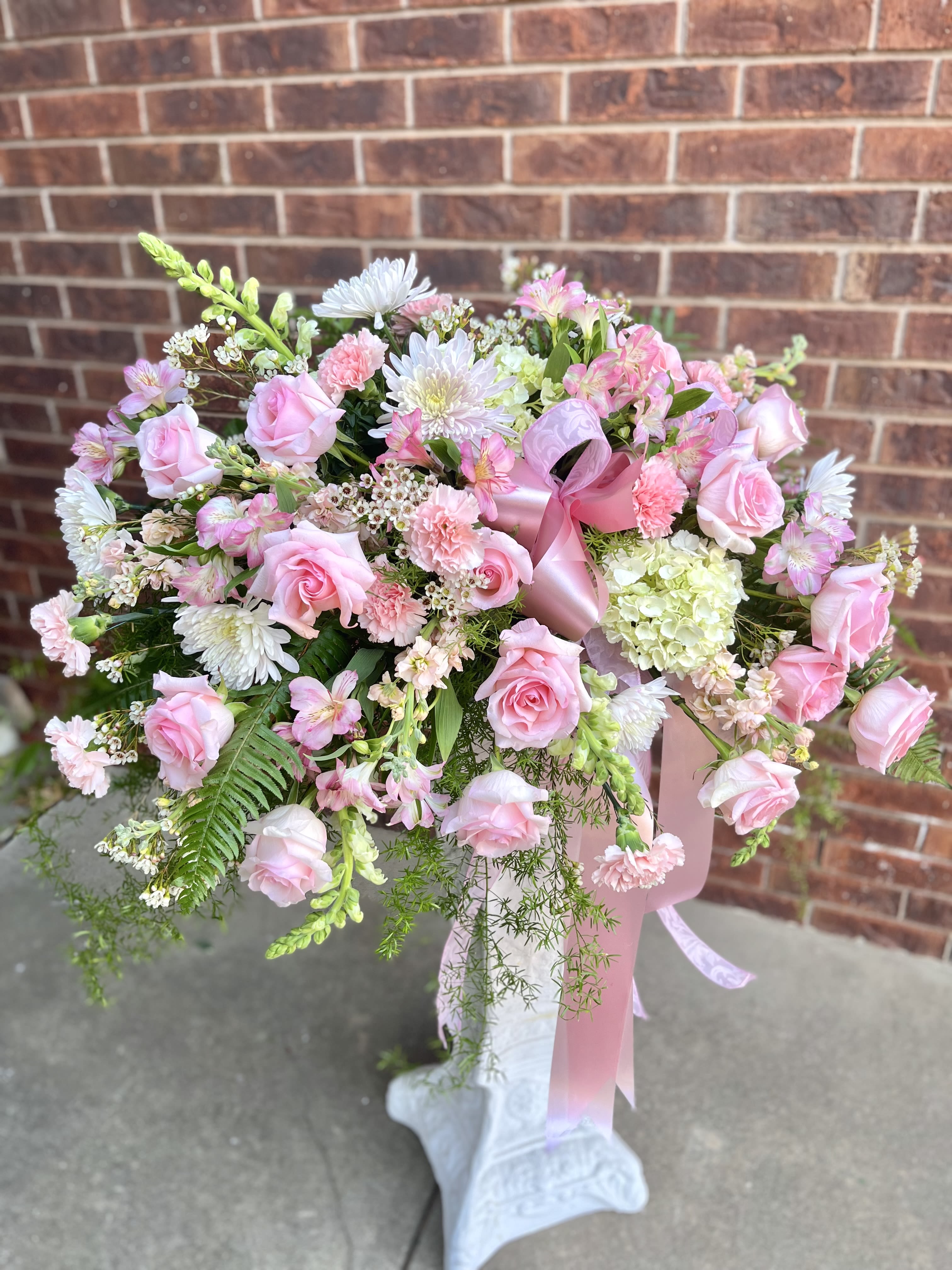 Embrace Tribute - A soft, garden-style pedestal arrangement in pink, white, and green that reads like a gentle embrace-perfect as a sympathy piece, wedding centerpiece, anniversary tribute, or Mother's Day surprise. This large, airy bouquet sits atop a white pedestal stand and fills the room with a romantic, feminine mood; the layered roses and daisy-like blooms give immediate visual impact while the overall spread reads as celebratory and memorable for the person you're honoring.  Carefully balanced textures-rounded roses, ruffled carnations, delicate alstroemeria and snapdragons, plus cheerful chrysanthemums and filler daisies-create a lush, layered look that's full from every angle. The design spreads broadly across the top of the pedestal, with a satin pink ribbon threaded through the center for an elegant finishing touch. Tones are soft pink and white warmed by fresh greenery for contrast.  Made to mark meaningful moments, this arrangement makes the recipient feel cherished and celebrated-ideal for mothers, brides, or a special anniversary partner. Delivered ready to display on its white pedestal stand; to keep blooms looking their best, place away from direct heat and refresh the water as needed.