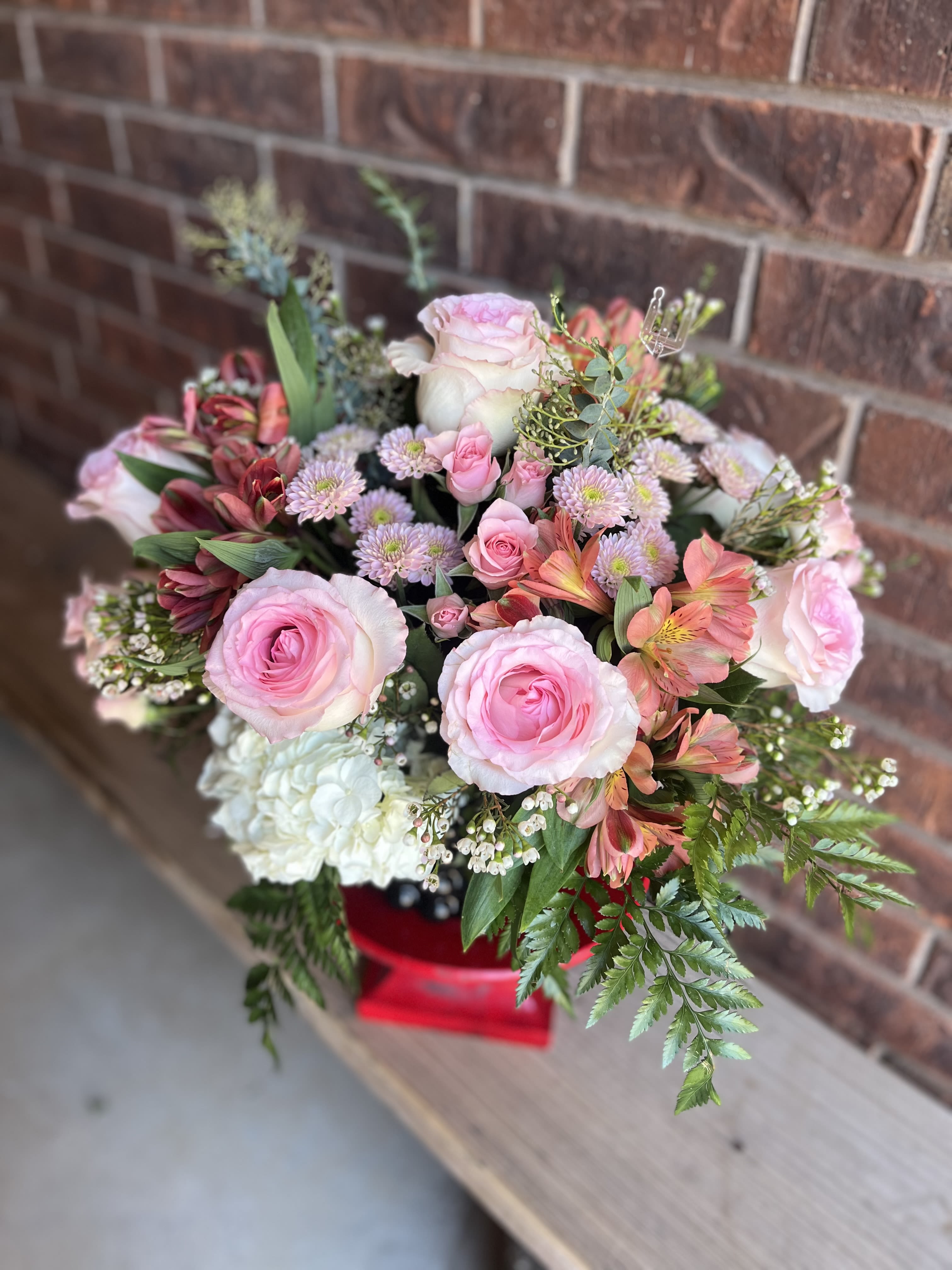 Daphne Rose - Large pink roses paired with coral alstroemeria and soft white chrysanthemums create a romantic, cheerful flower arrangement perfect as a birthday bouquet, anniversary roses alternative, or heartfelt gift for someone you cherish. The rounded, full design highlights open rose petals and smaller spray roses for depth, while fern-like foliage and clustered filler blooms add a textured, garden-fresh feel that makes the recipient feel truly seen.  This arrangement features large pink roses with smaller pink spray roses, white chrysanthemum blooms, and coral alstroemeria in a lush, layered composition. A beautiful ceramic container anchors the display, giving a striking contrast to the pastel palette and making the presentation feel festive and gift-ready. The mix of pink, white, red, and mixed tones creates a versatile look that reads both romantic and celebratory.  Choose this as a meaningful way to say "happy birthday," "congratulations," or "I'm thinking of you" - she'll feel uplifted by the color and texture of the blooms. Arrangement is delivered in the red gift box shown; for best longevity, keep flowers in a cool spot away from direct sun and refresh the water if provided instructions accompany the delivery.