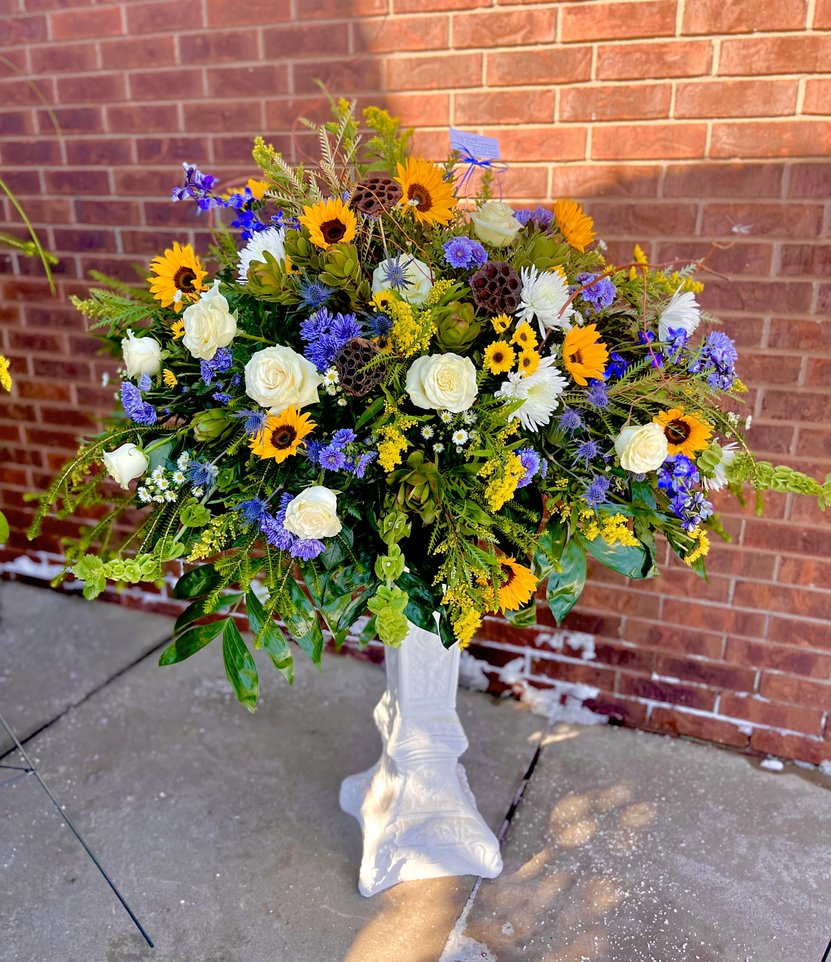 Rustic Sun Tribute - A burst of sunshine and quiet reverence, this flower arrangement pairs bold sunflowers with creamy white roses for a Rustic Sun Tribute that suits both celebratory and memorial occasions. As a sympathy arrangement or a bright centerpiece for a celebration of life, the mixed-color palette of yellow, white, and purple reads hopeful and uplifting-designed to make the person you're honoring feel seen and remembered.  The design features large yellow sunflowers, soft white roses, white daisies, and clusters of purple aster and other seasonal fillers arranged in a full, rounded, garden-style silhouette. Tall, airy stems and varied textures give the bouquet a loose, abundant look while the white pedestal vase lends a formal, elevated presentation and keeps the display clean and ceremonial. The overall scale is large and generous, ideal for making a meaningful visual impact in a room or at a service.  This arrangement is a thoughtful way to celebrate a life, mark a sympathy moment, or bring warm, rustic cheer to a home. The combination of hardy sunflowers and classic roses keeps blooms looking fresh longer; place in a cool spot away from direct heat and refresh the water daily to extend vase life. Order with confidence for delivery to a service, memorial, or to send a comforting, beautiful message to someone special.