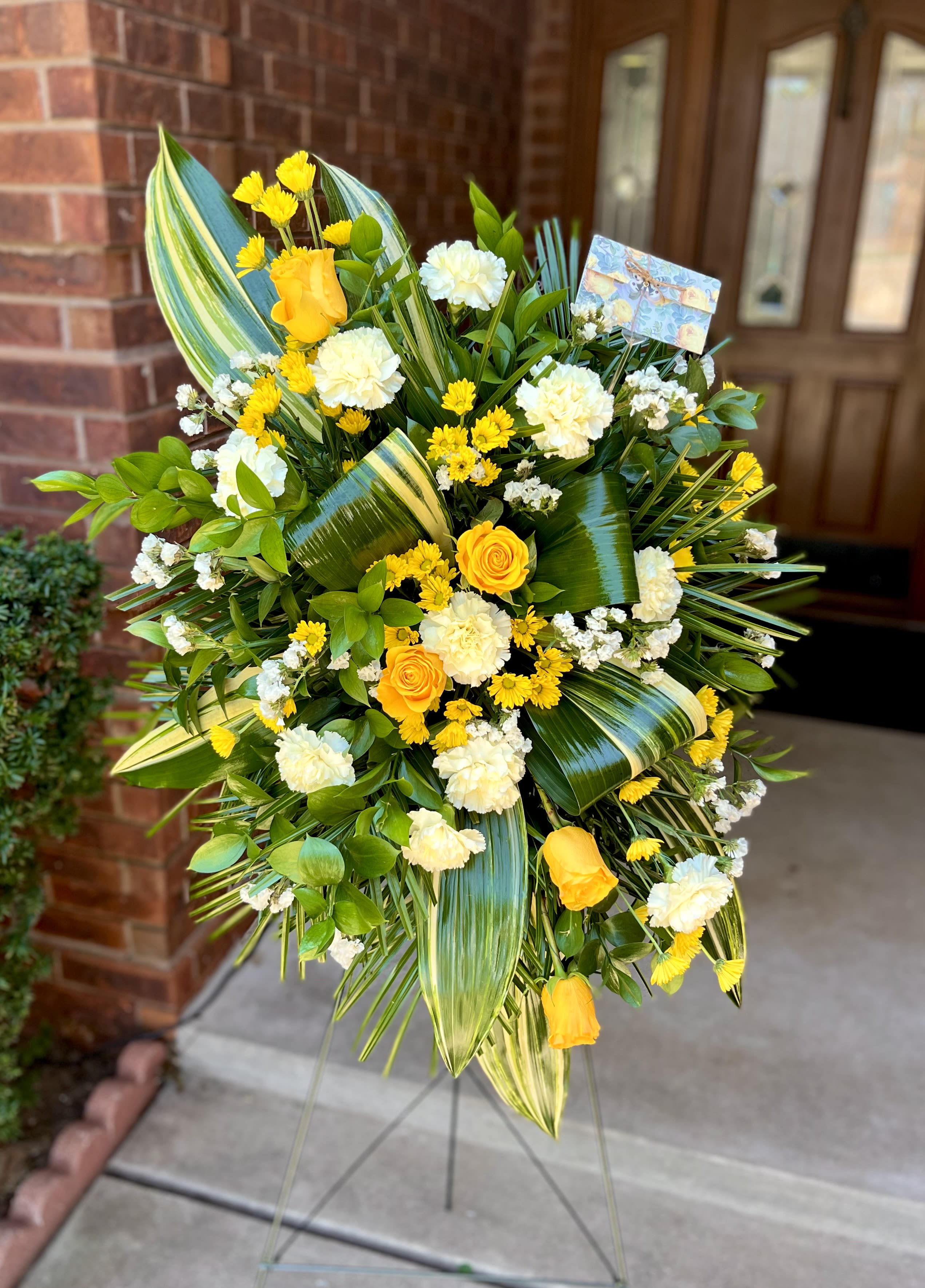 Golden Grace - Sunny yellow roses set against crisp white carnations give this standing spray an uplifting glow - ideal as a sympathy arrangement or memorial tribute that feels warm without being flashy. The rounded, full shape and tropical, striped palm fronds create a bold green frame so the recipient will feel comforted and gently uplifted during a difficult moment.  This flower arrangement includes yellow roses, white carnations, yellow daisy-like chrysanthemums and layered greenery arranged on a standing easel; a small card is tucked into the upper right for your personal message. The palette of yellow, white, and green reads fresh and formal, with palm fronds and foliage adding texture and a statuesque silhouette appropriate for services and remembrance.  Choose this design when you want the person you're honoring to be remembered with brightness and dignity - a thoughtful way to say you're thinking of them. The spray arrives ready to display on its standing easel; include your message on the provided card when placing your order.