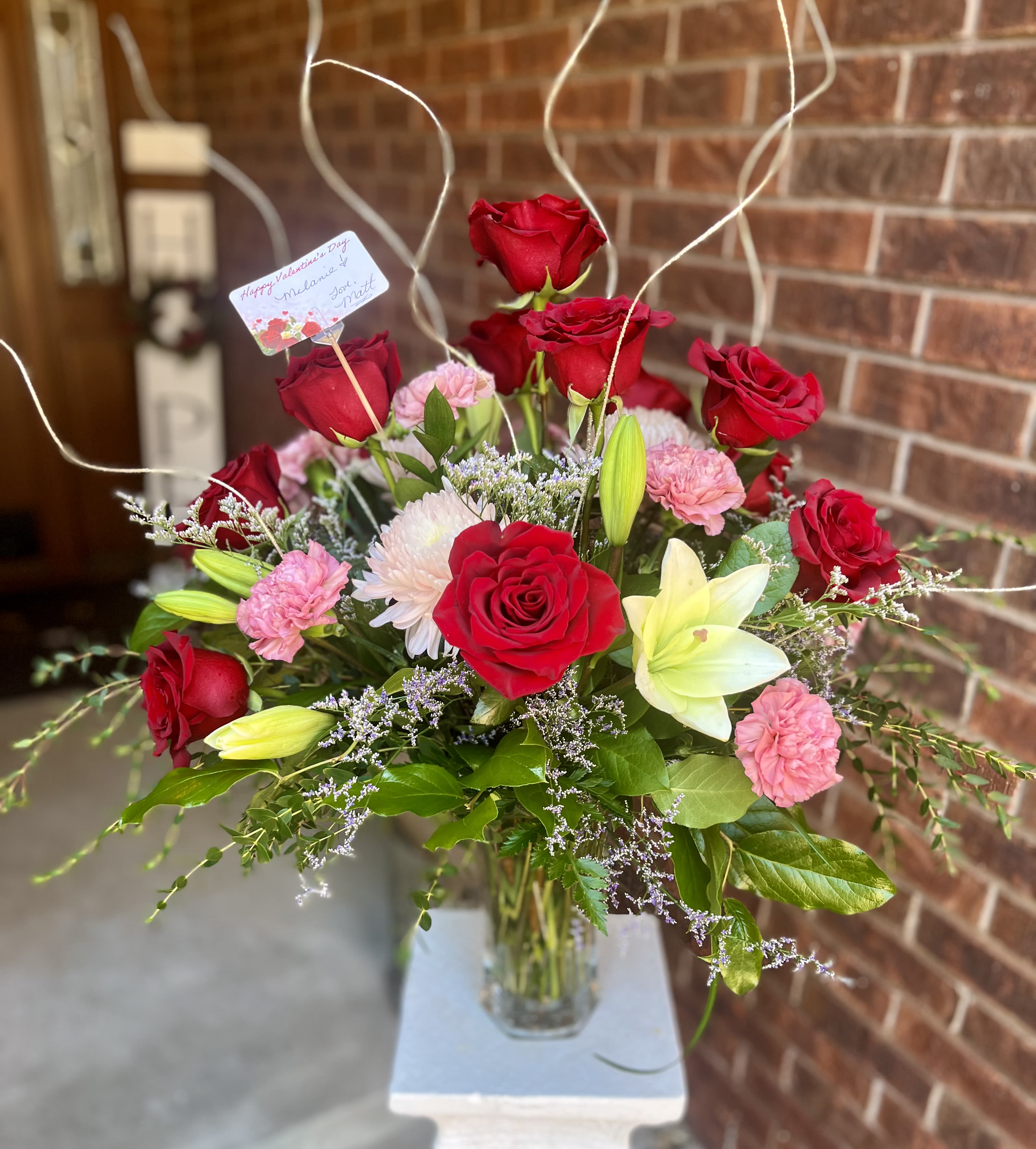 Mixed Dozen - A clustered dozen mixed bouquet of roses, carnations and lilies that reads romantic and cheerful-the perfect birthday or Valentine's Day bouquet to make someone feel adored. The arrangement's dominant red roses and soft pink carnations are lifted by pale white blooms and a single light yellow lily, creating a warm, layered palette that says "you matter" without being fussy.  Arranged in a clear glass vase, glossy green foliage and fine airy filler give the bouquet depth and a soft, textured silhouette. Includes a small card on a wooden pick for your personal message; the vase provides instant display so the recipient can enjoy the scent and color immediately. Ideal for anniversaries, sweetheart gestures, or to celebrate a special friend-sizes and care details are shown at checkout for easy ordering and freshness guidance.